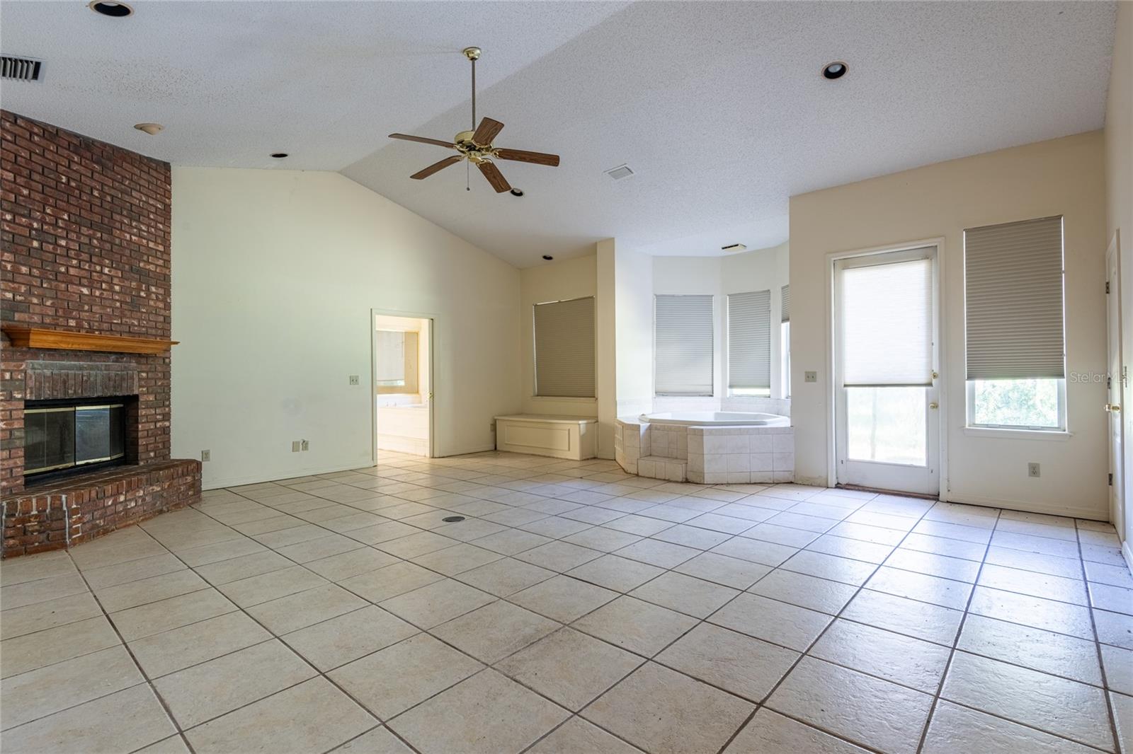 Empty tiled living area with a brick fireplace, ceiling fan, and corner soaking tub.