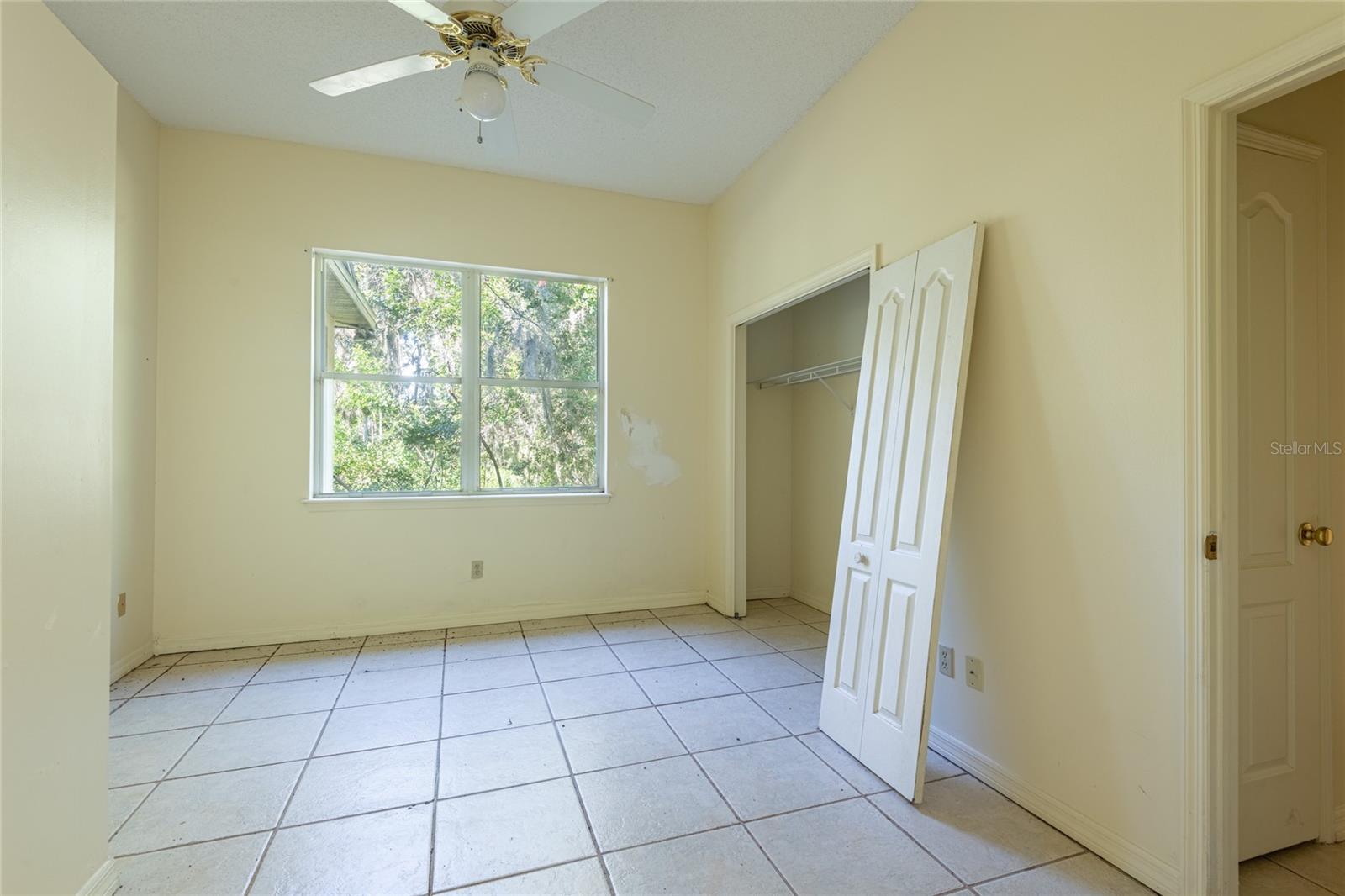 Empty bedroom with tiled floor, ceiling fan, tree-facing window, and open closet.