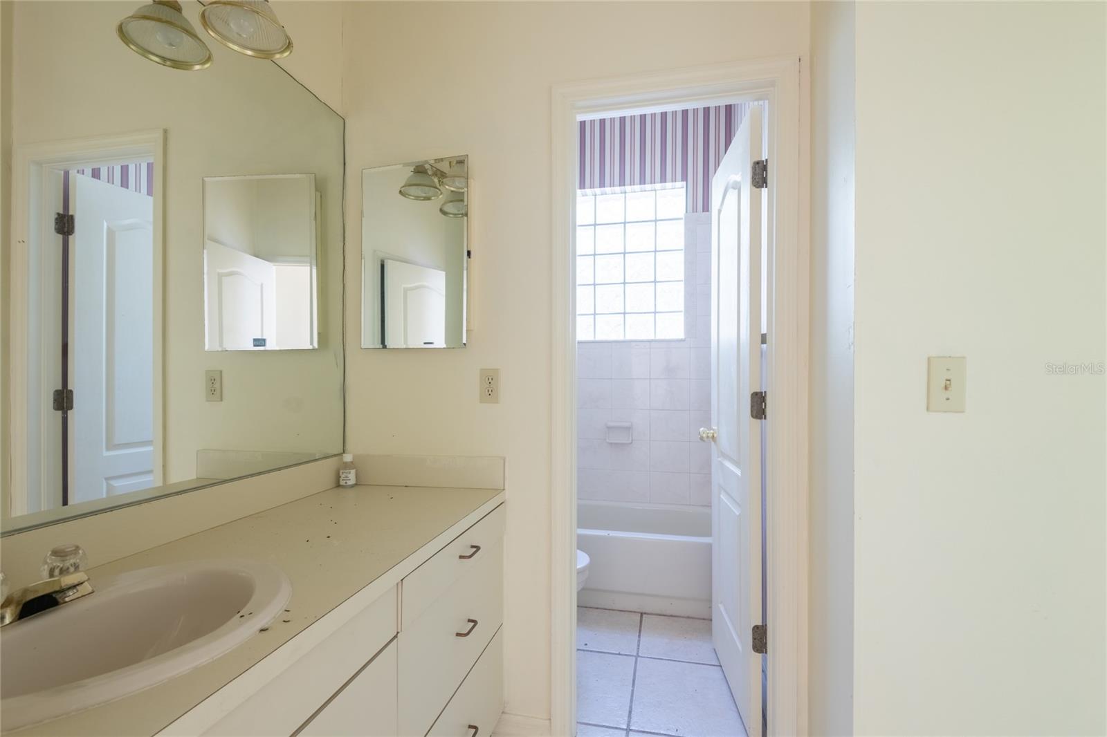 Bathroom with a long white vanity and mirrors opening onto a tiled tub and shower.