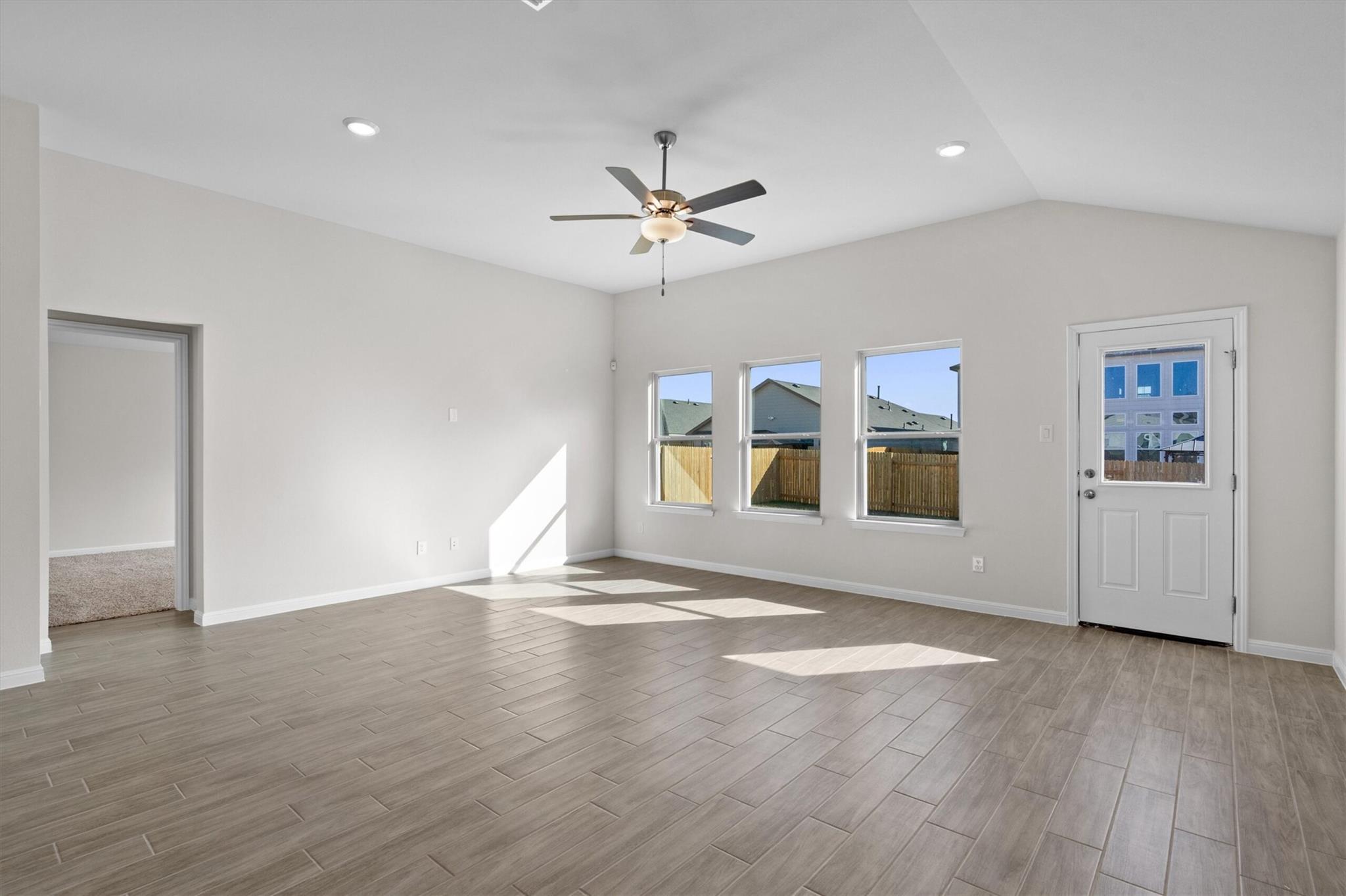 Empty living room with tile floors, vaulted ceiling, ceiling fan, three windows and a glass-panel door.