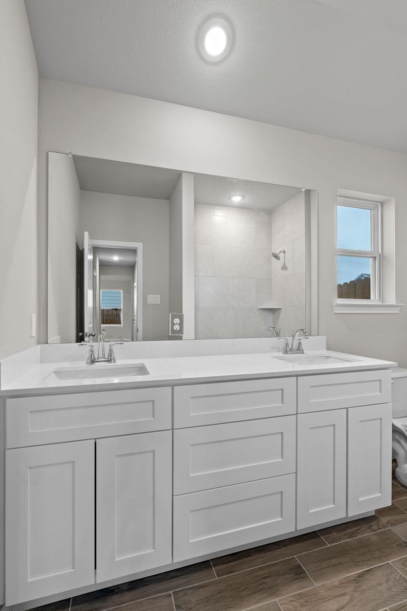 Bathroom with a white double-sink vanity, large mirror, tiled shower, and dark wood-look flooring.