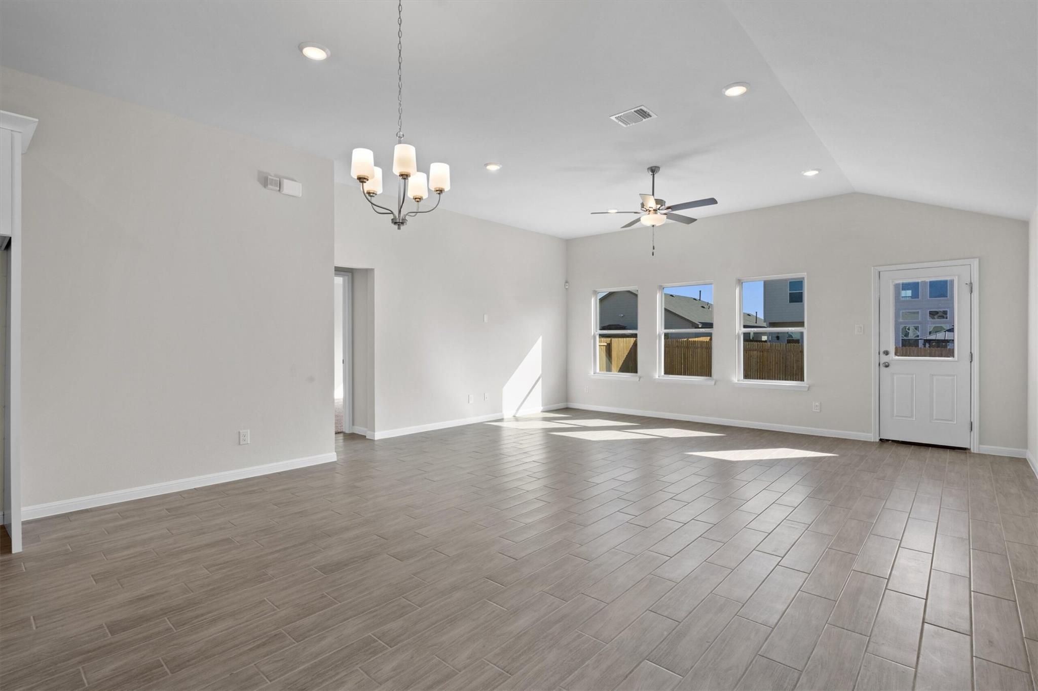 Empty living and dining area with gray tile floors, chandelier, ceiling fan, and three windows.