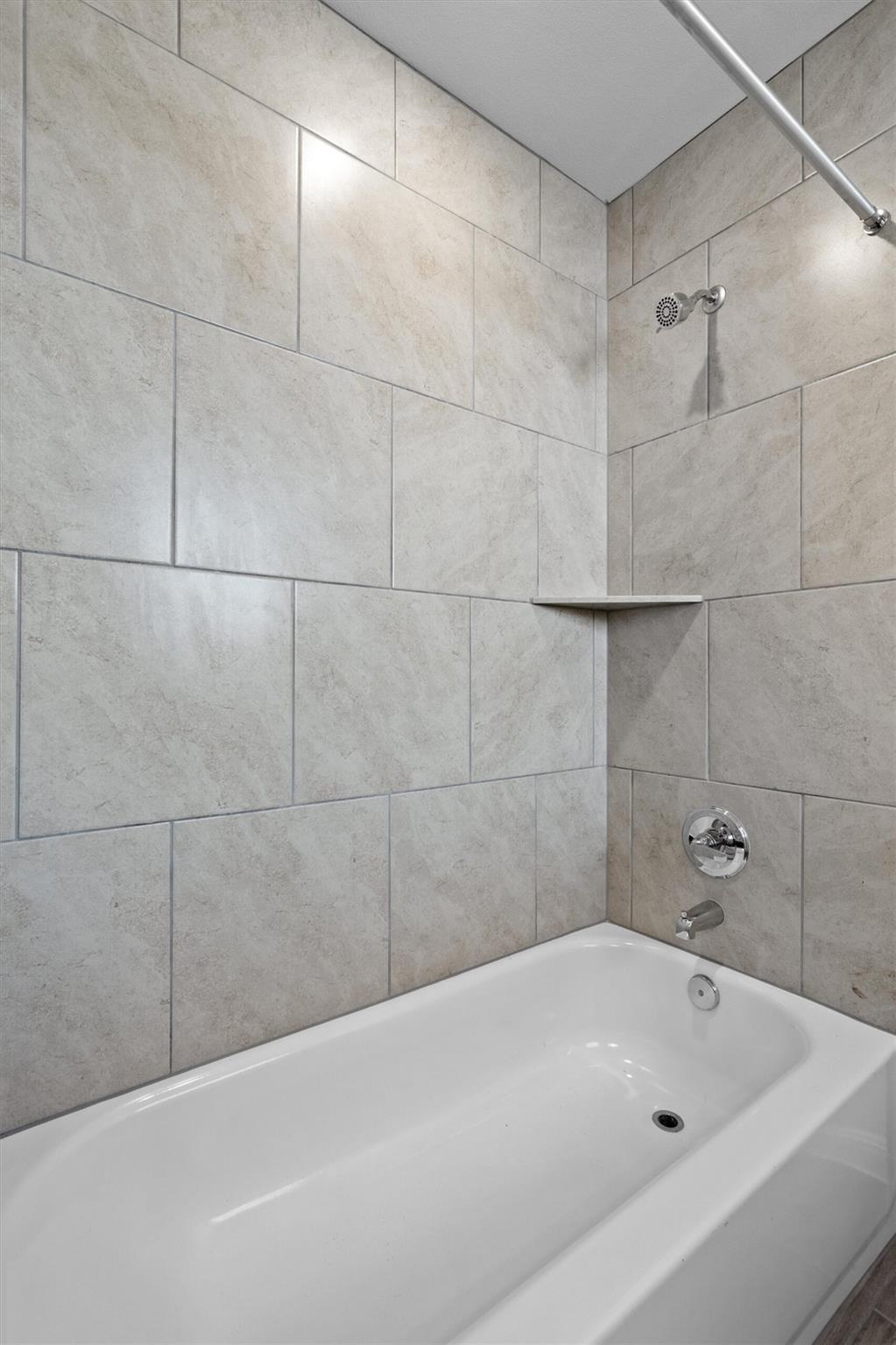 White bathtub in a tiled shower with a corner shelf and chrome fixtures.