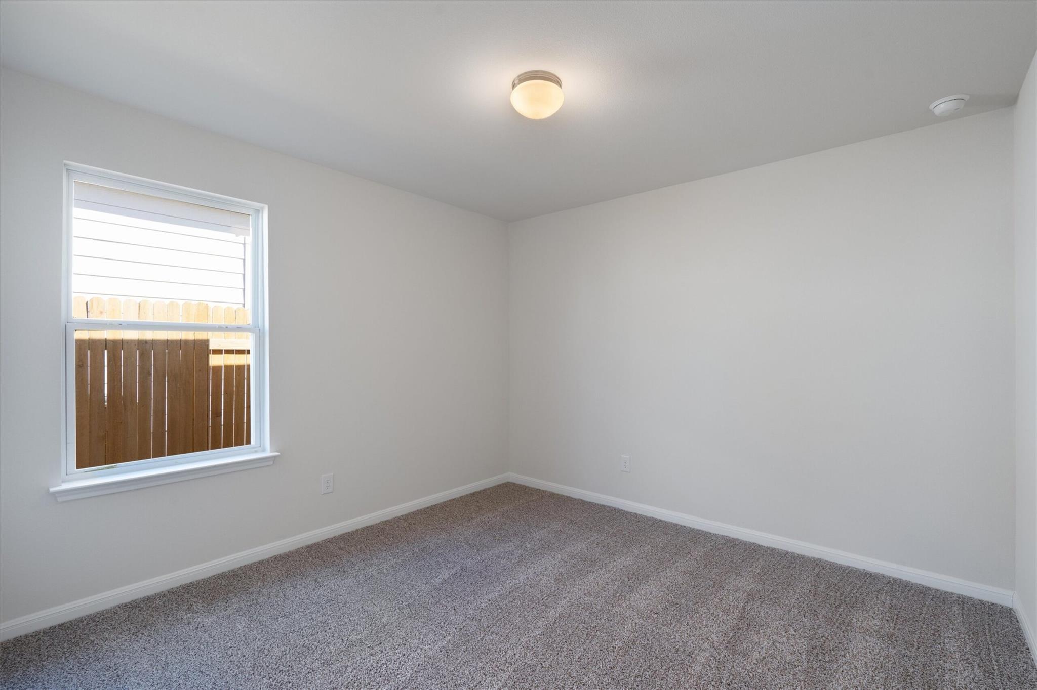 Empty carpeted room with gray walls and a window overlooking a wooden fence.