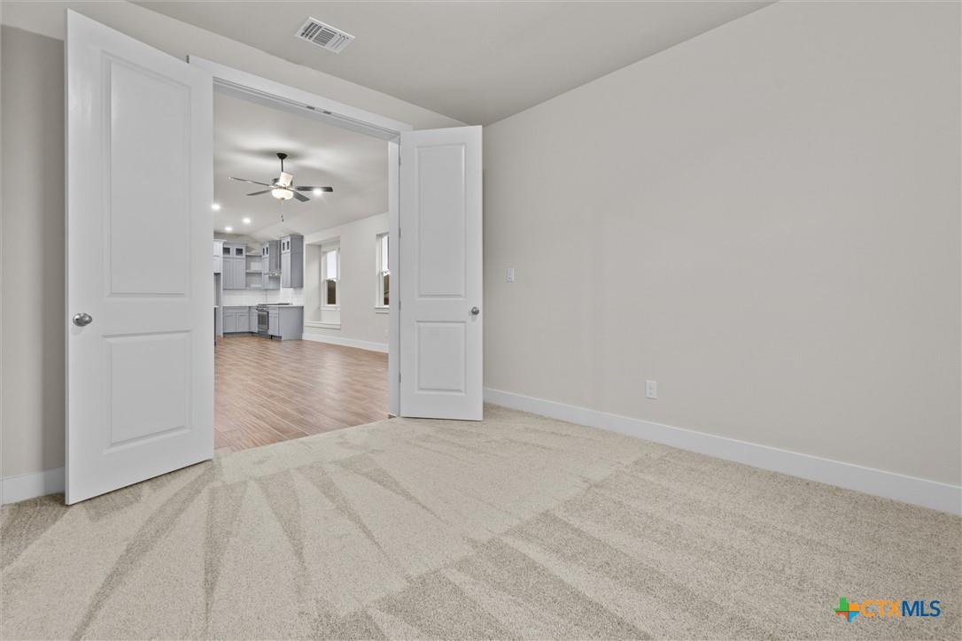 Empty carpeted room with open double doors leading to a kitchen.
