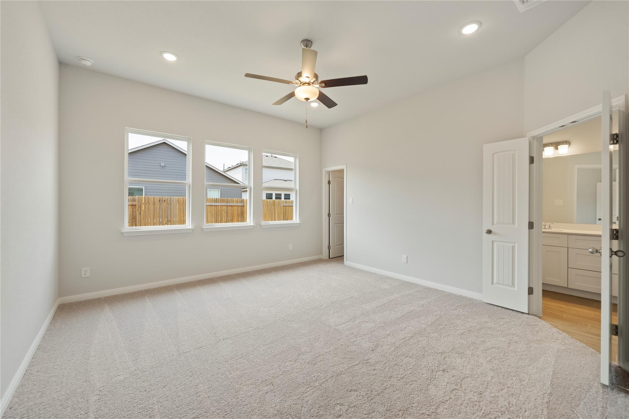 Empty carpeted bedroom with three windows, a ceiling fan, and an ensuite bathroom doorway.
