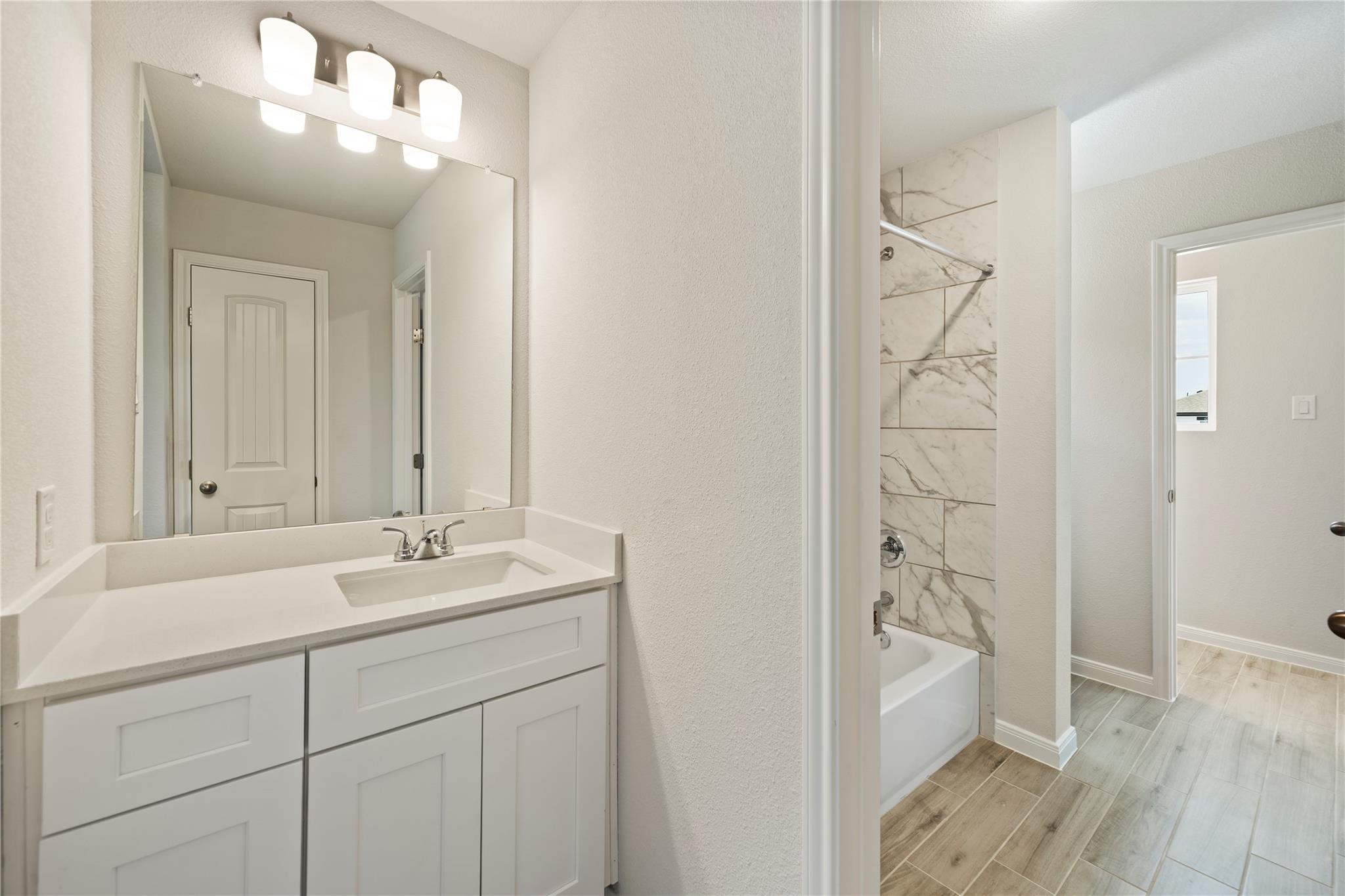 Bathroom vanity and sink beside a bathtub with marble-look tile surround and wood-look flooring.