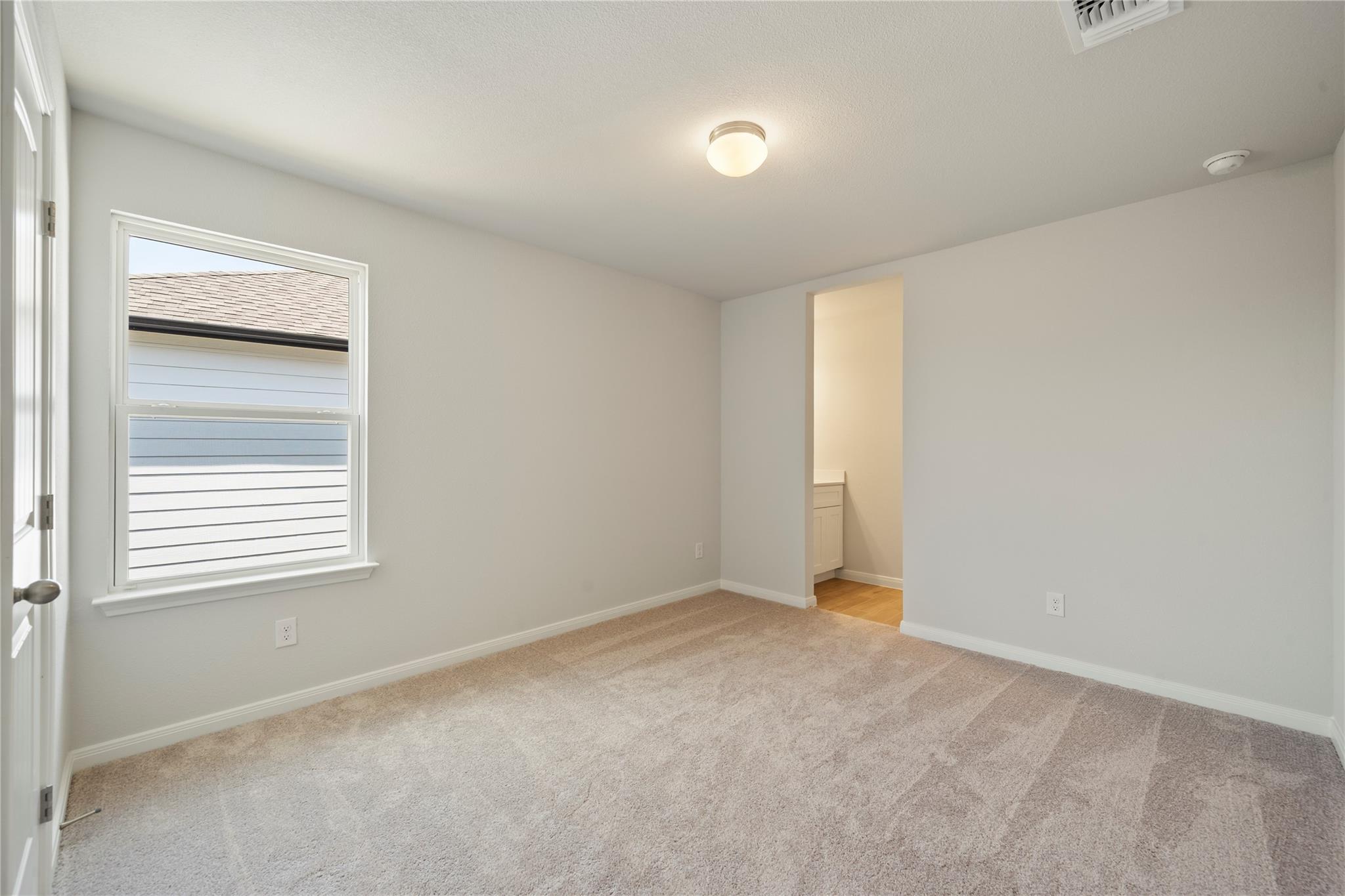 Empty carpeted bedroom with a window and an open doorway to a bathroom vanity.