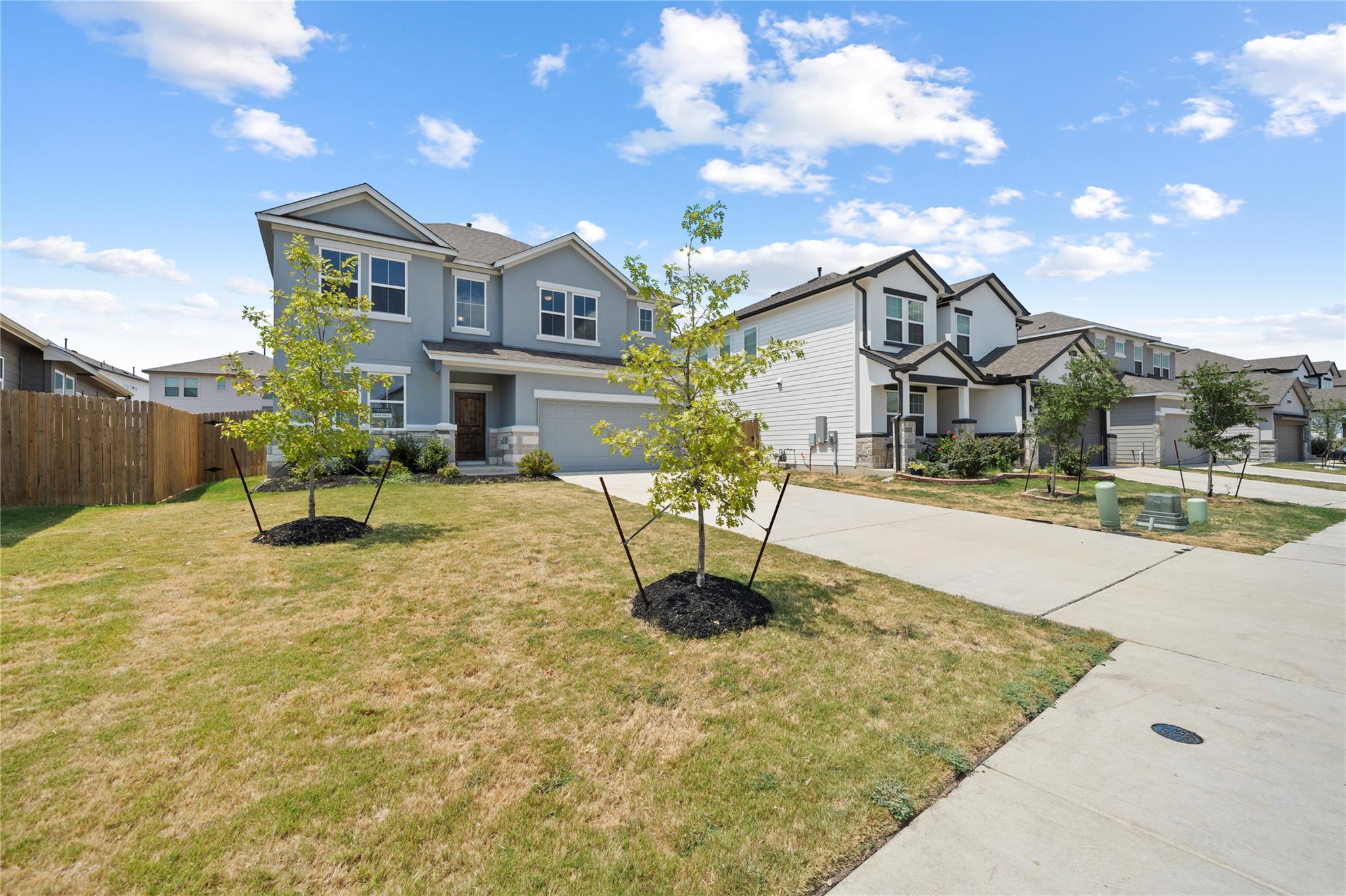Two-story blue-gray house with a two-car garage, stone accents, and young trees in the front yard.