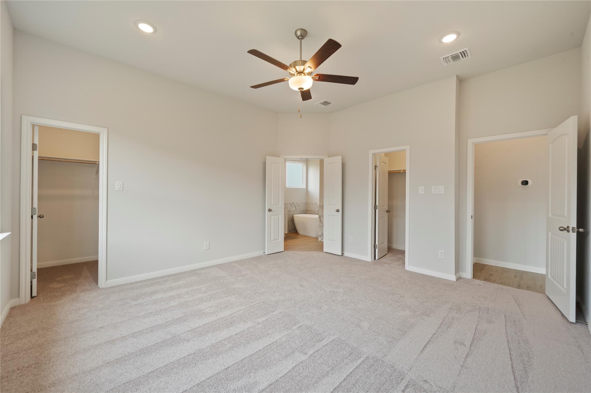 Empty carpeted bedroom with a ceiling fan and open doors to closets and a bathroom.