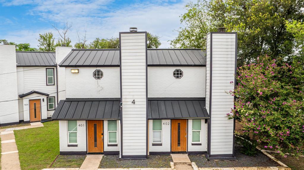 White multi-unit building with dark metal awnings, orange-brown doors, and flowering shrubs.