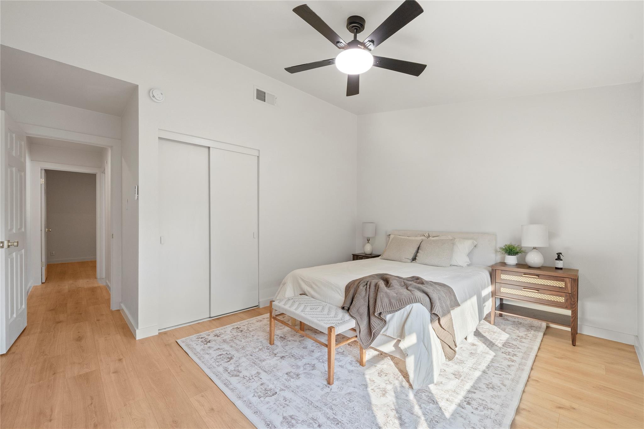 Bright bedroom with a white bed, patterned rug, sliding closet doors and dark ceiling fan.