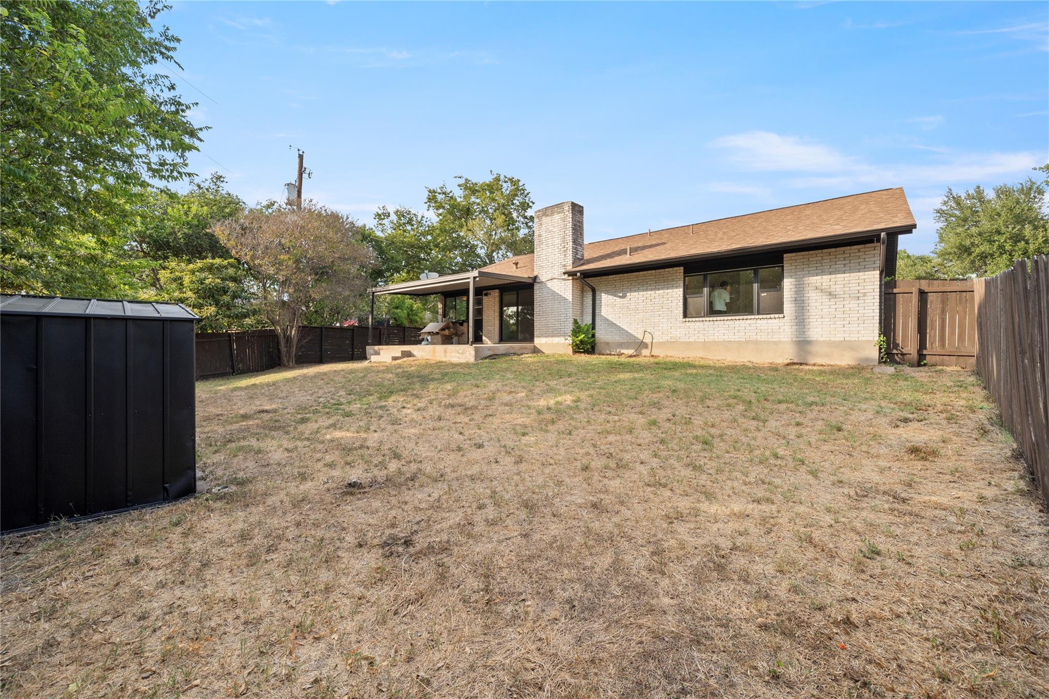 Rear of a light brick house with a covered patio, fenced yard, and black storage shed.