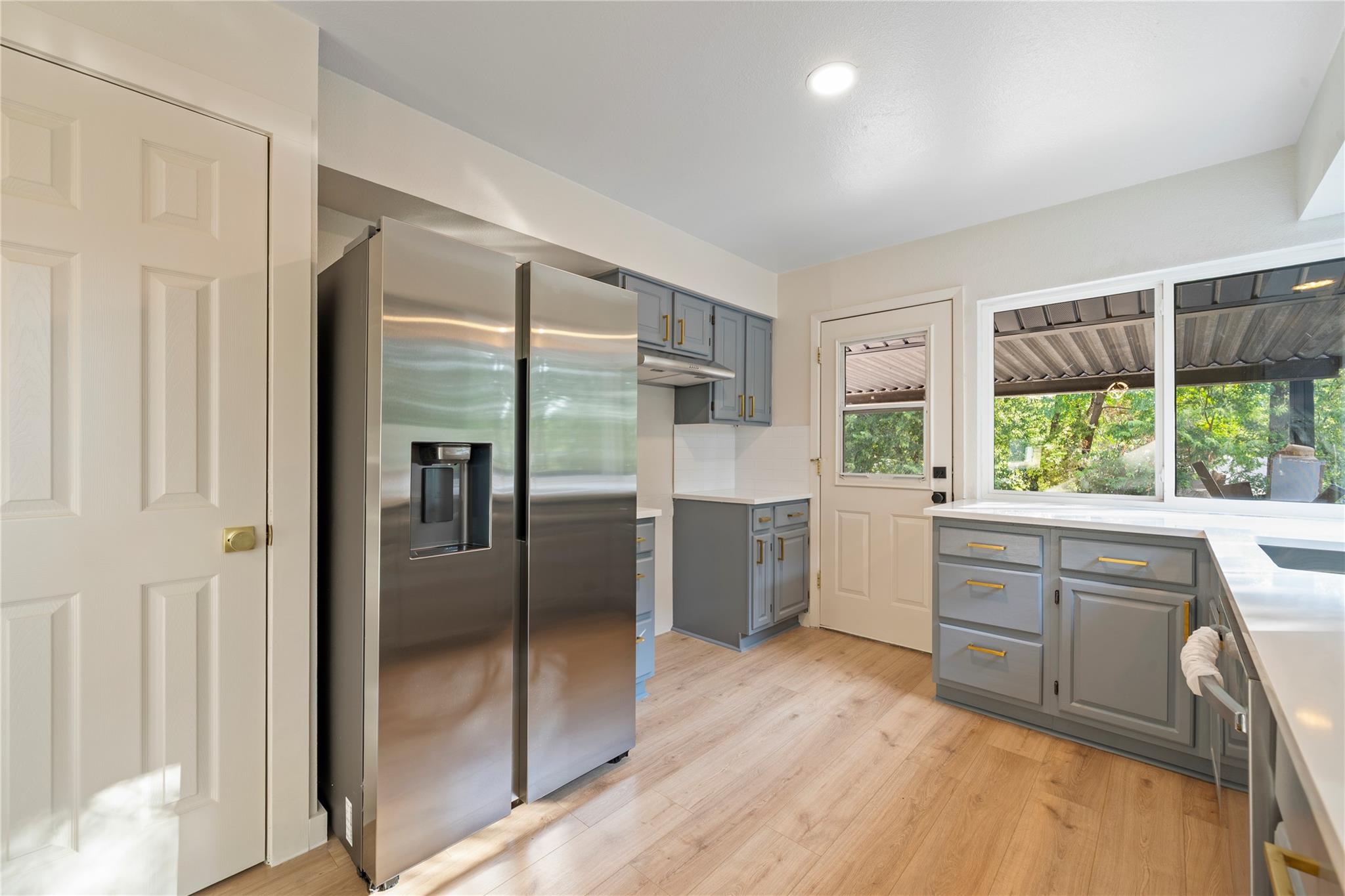 Kitchen with gray cabinets and gold pulls, stainless steel refrigerator, and a large window overlooking greenery.