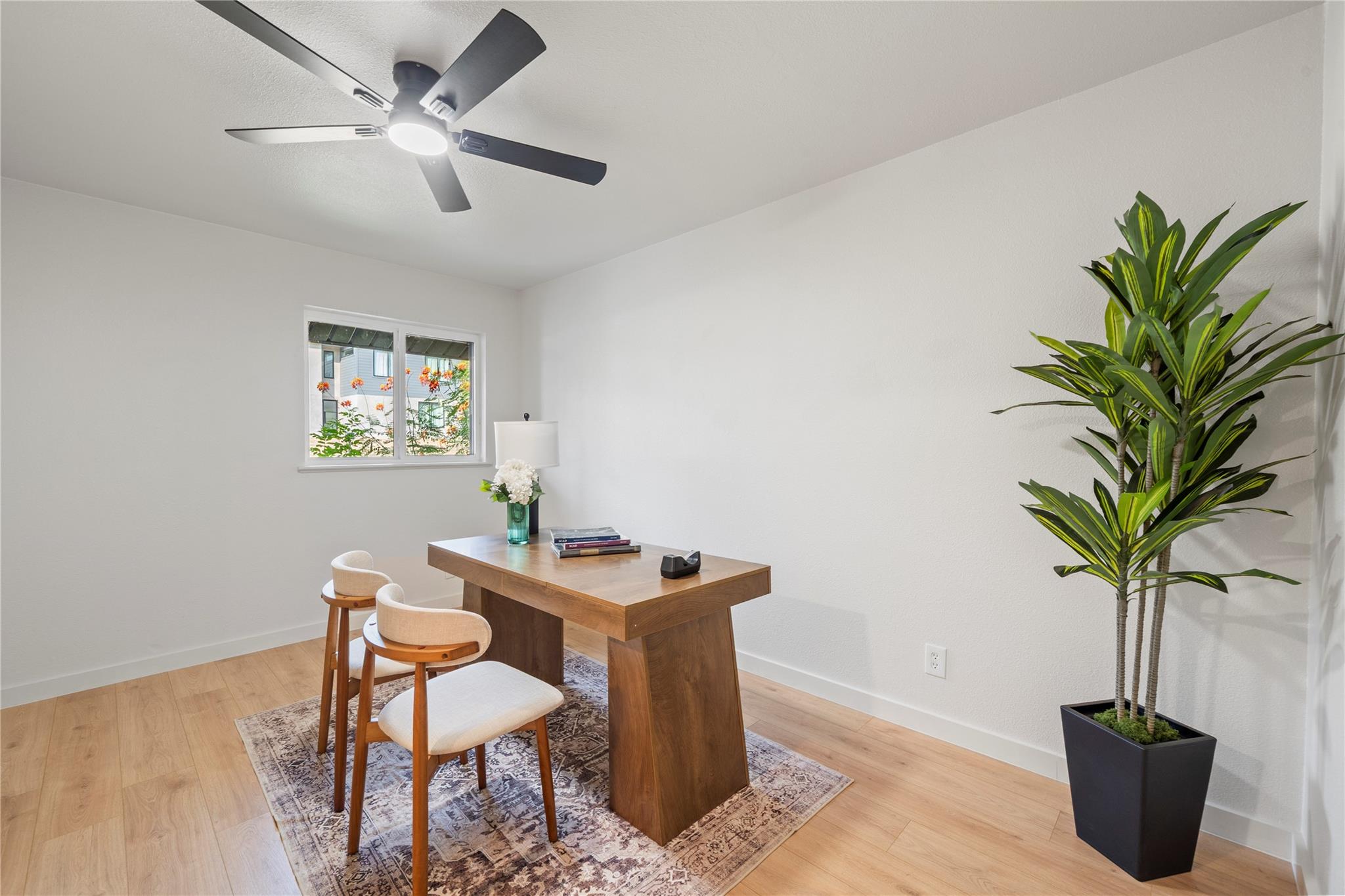 Staged home office with a wooden desk, two chairs, patterned rug, and tall potted plant.