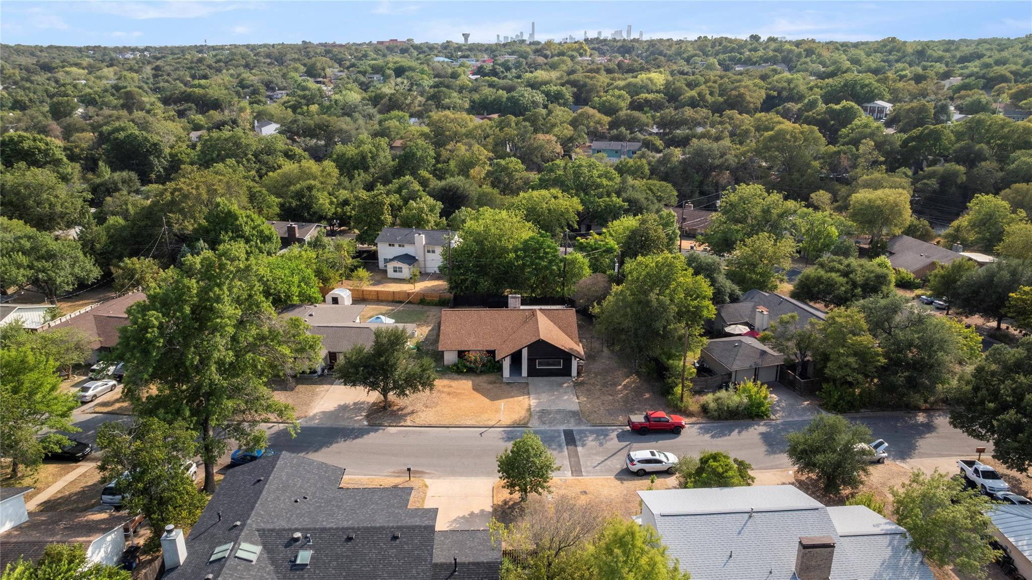 Aerial view of a dark Austin home with a brown roof, surrounded by trees and the distant downtown skyline.