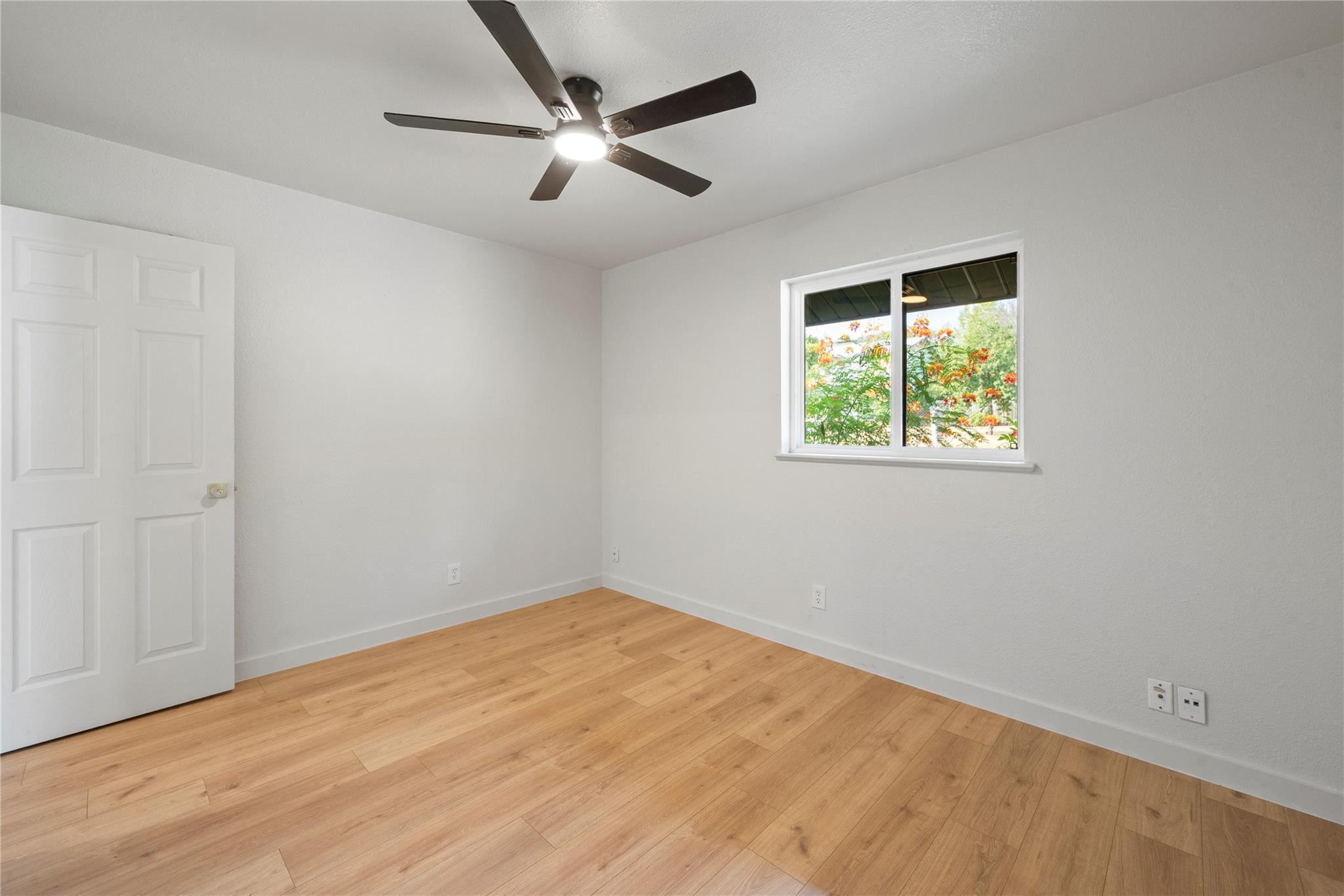 Empty room with light wood floors, a dark ceiling fan, and a window overlooking flowering trees.