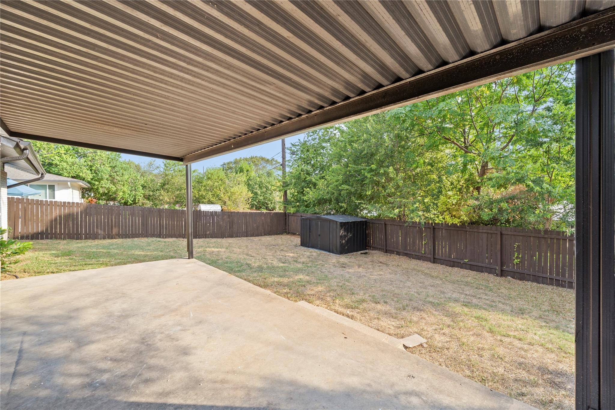 Covered patio overlooking a fenced backyard with a storage shed and mature trees.