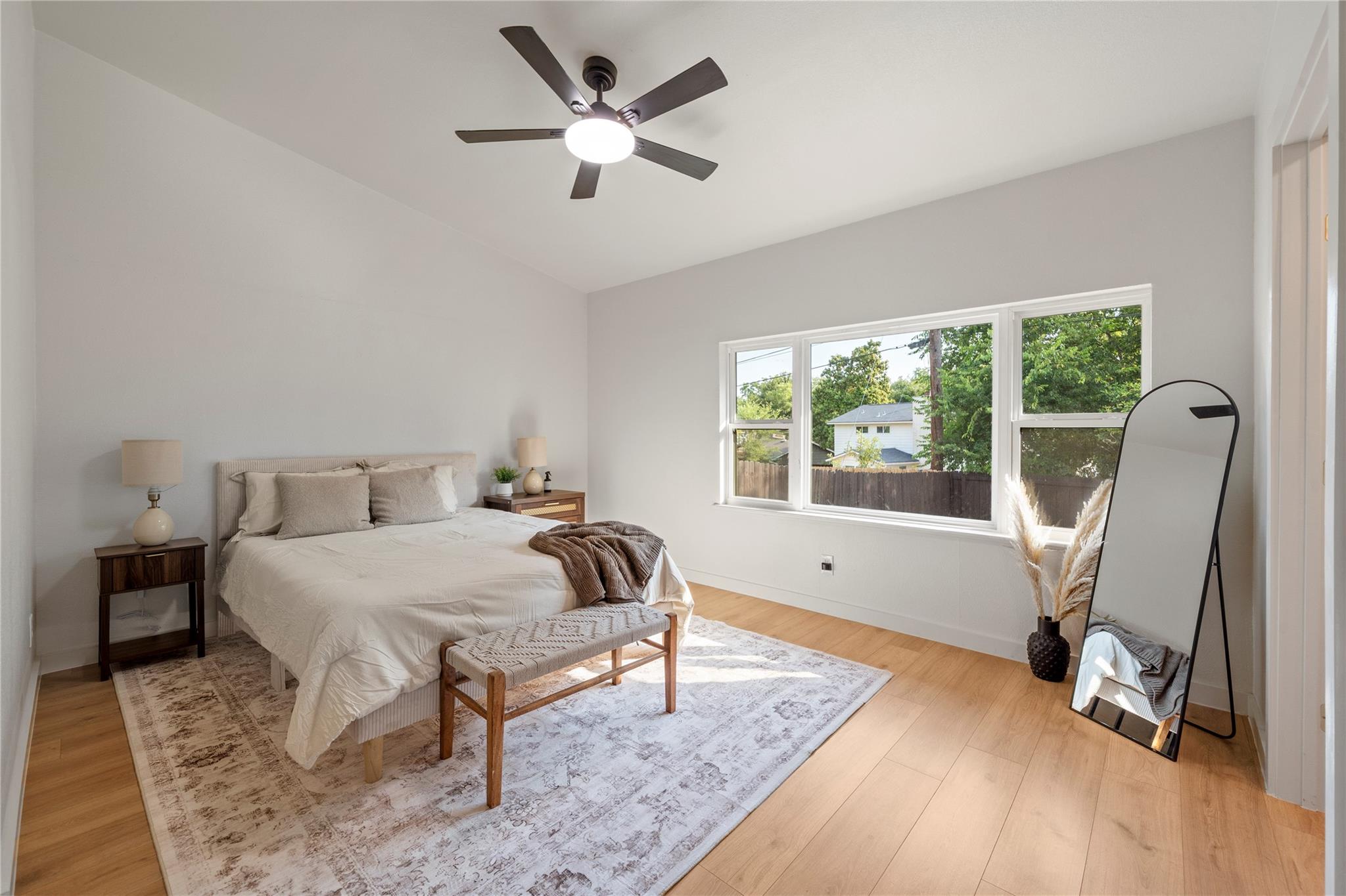 Furnished bedroom with a king-size bed, large windows, light wood floors, and a ceiling fan.