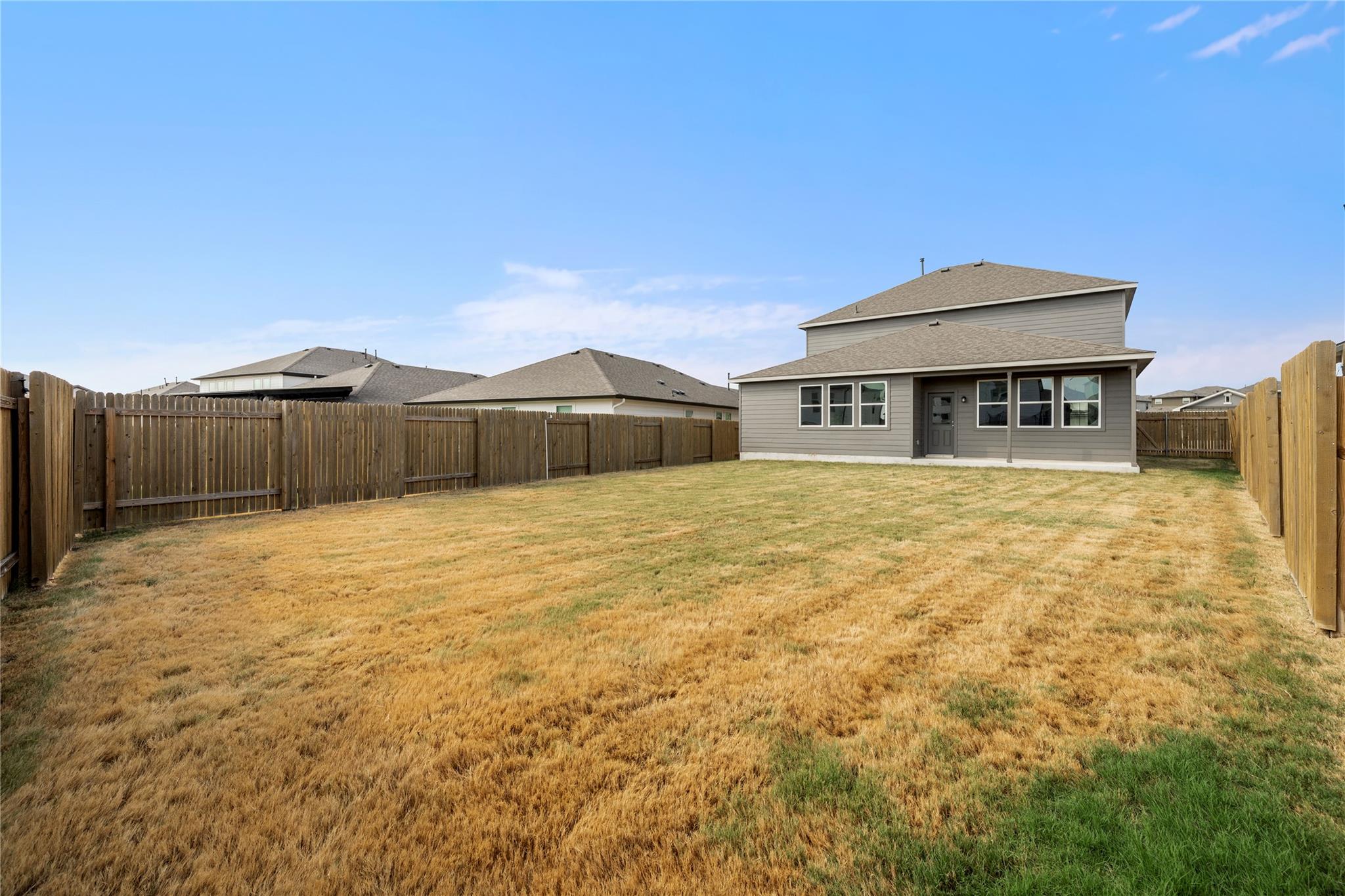 Two-story gray home behind a fenced backyard with a broad, dry lawn.