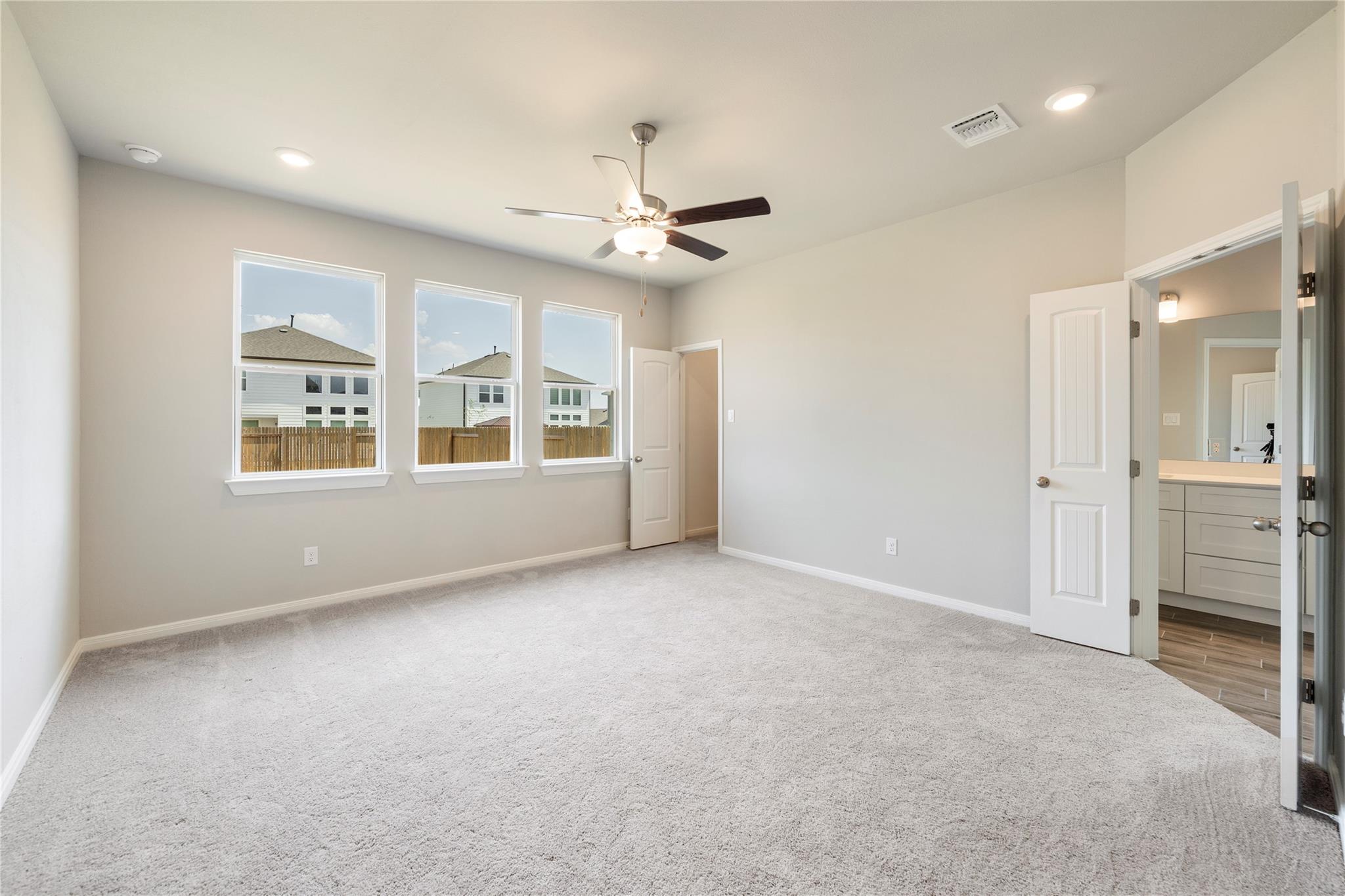 Vacant bedroom with three windows, light carpet, a ceiling fan, and an open doorway to the ensuite bathroom.