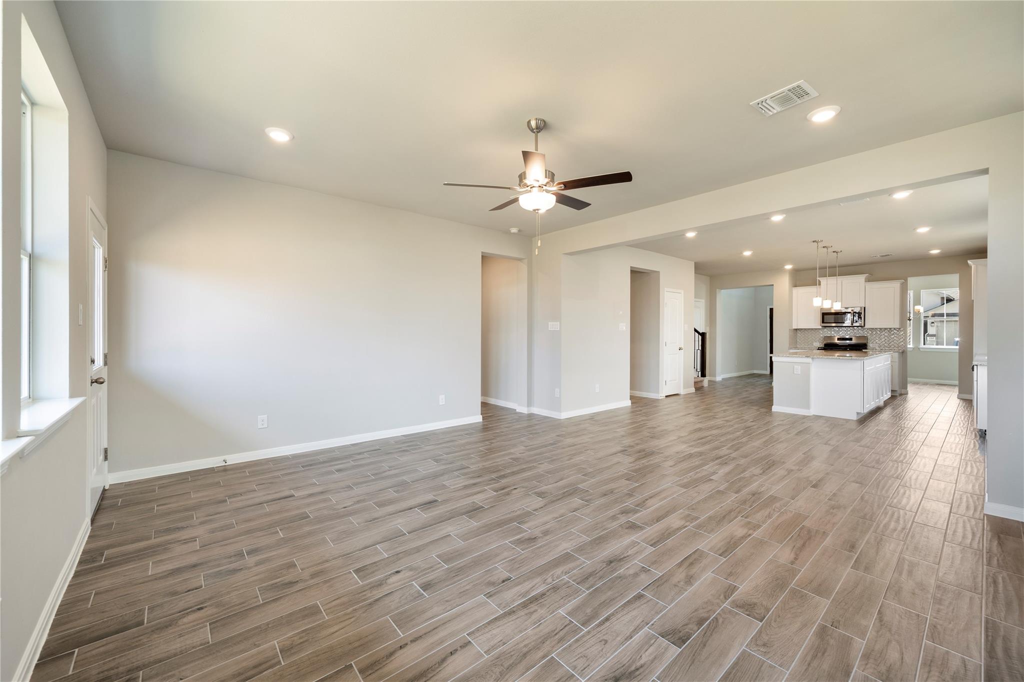 Open-plan living room and kitchen with wood-look tile floors, white island, and ceiling fan.