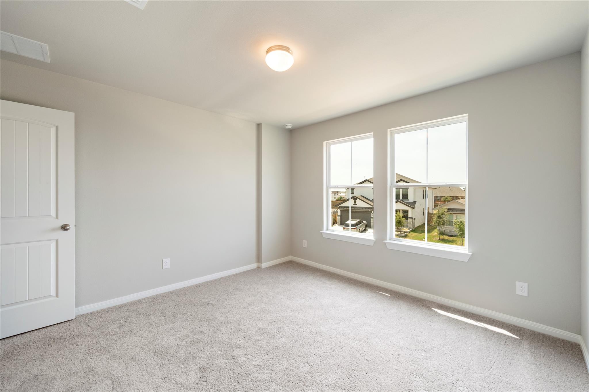 Empty bedroom with beige carpet and two windows overlooking neighboring homes.