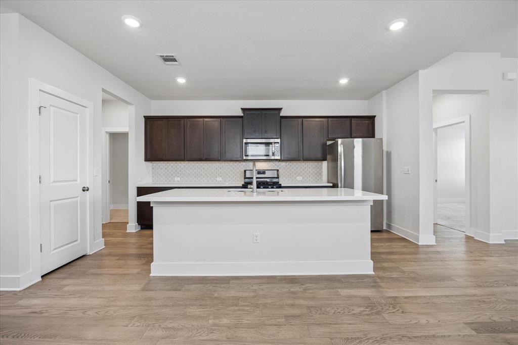 Kitchen with a large white island, dark cabinetry, stainless steel appliances, and wood-look flooring.