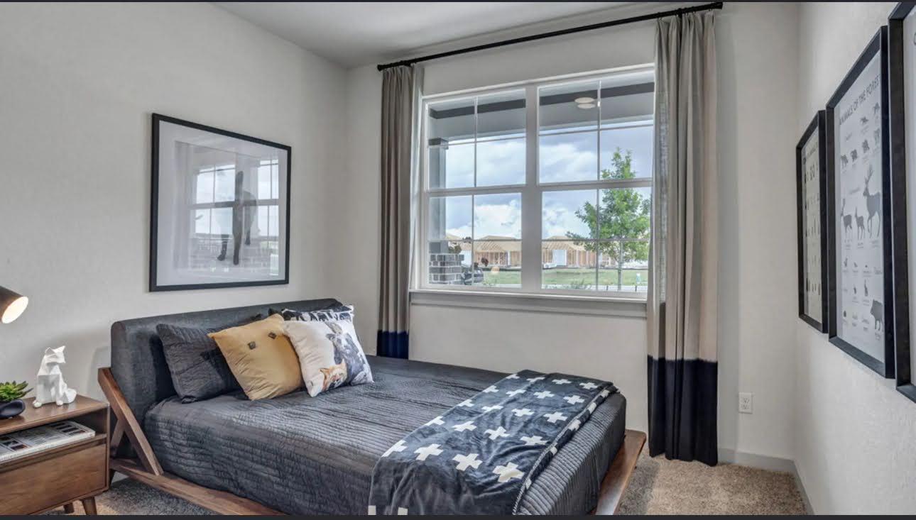 Bedroom with a gray bed and patterned blanket beside a large window overlooking neighboring homes.