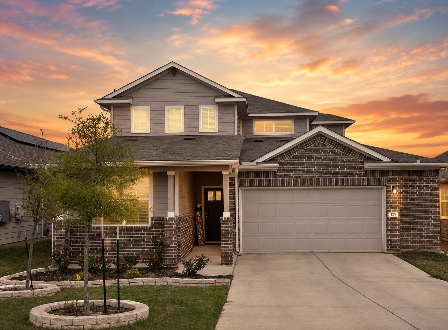 Two-story gray and brick house with a double garage, landscaped yard, and sunset sky.