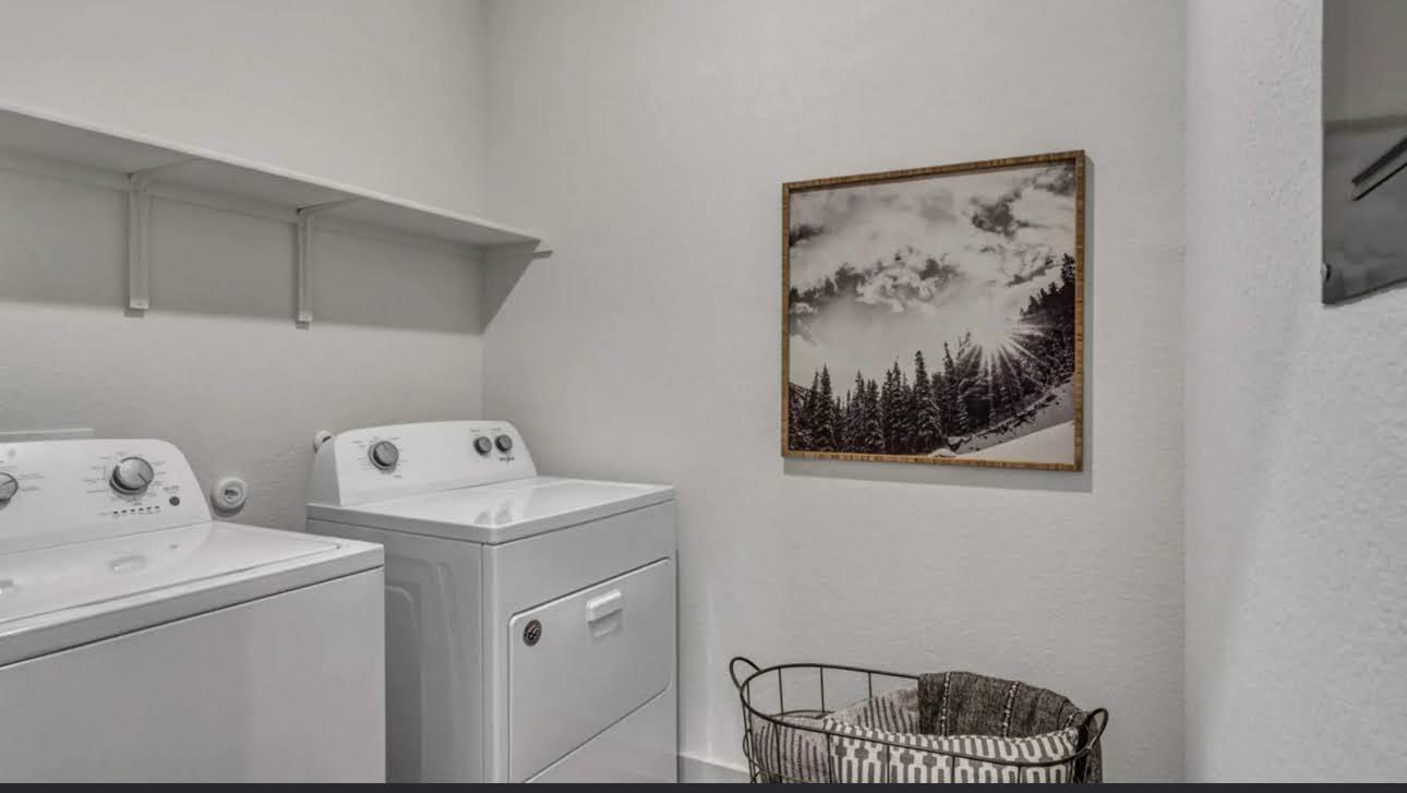 Laundry room with white washer and dryer beneath a shelf, with framed forest artwork and a laundry basket.
