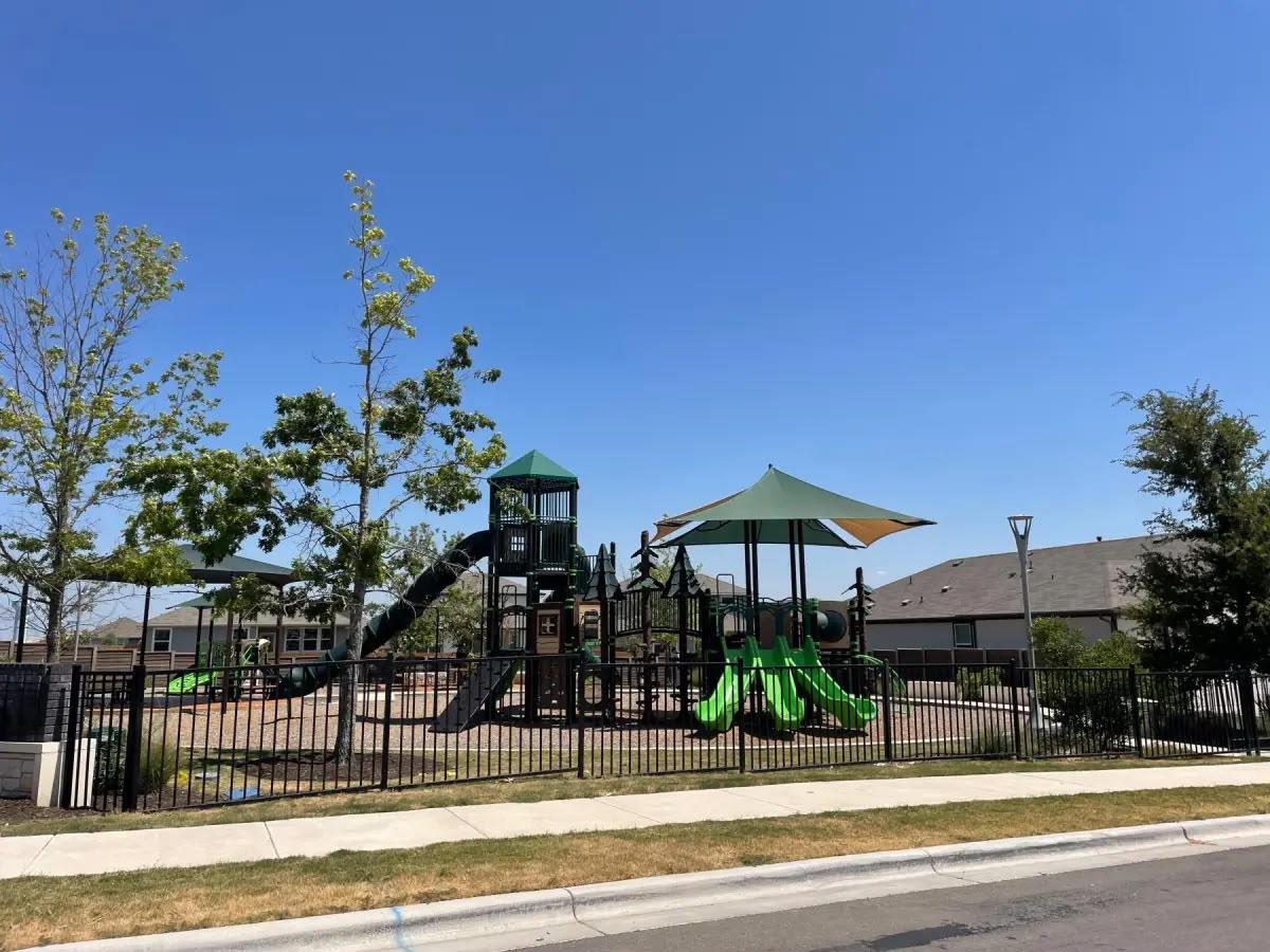 Neighborhood playground with green slides, climbing structures, and a shade canopy behind a black fence.