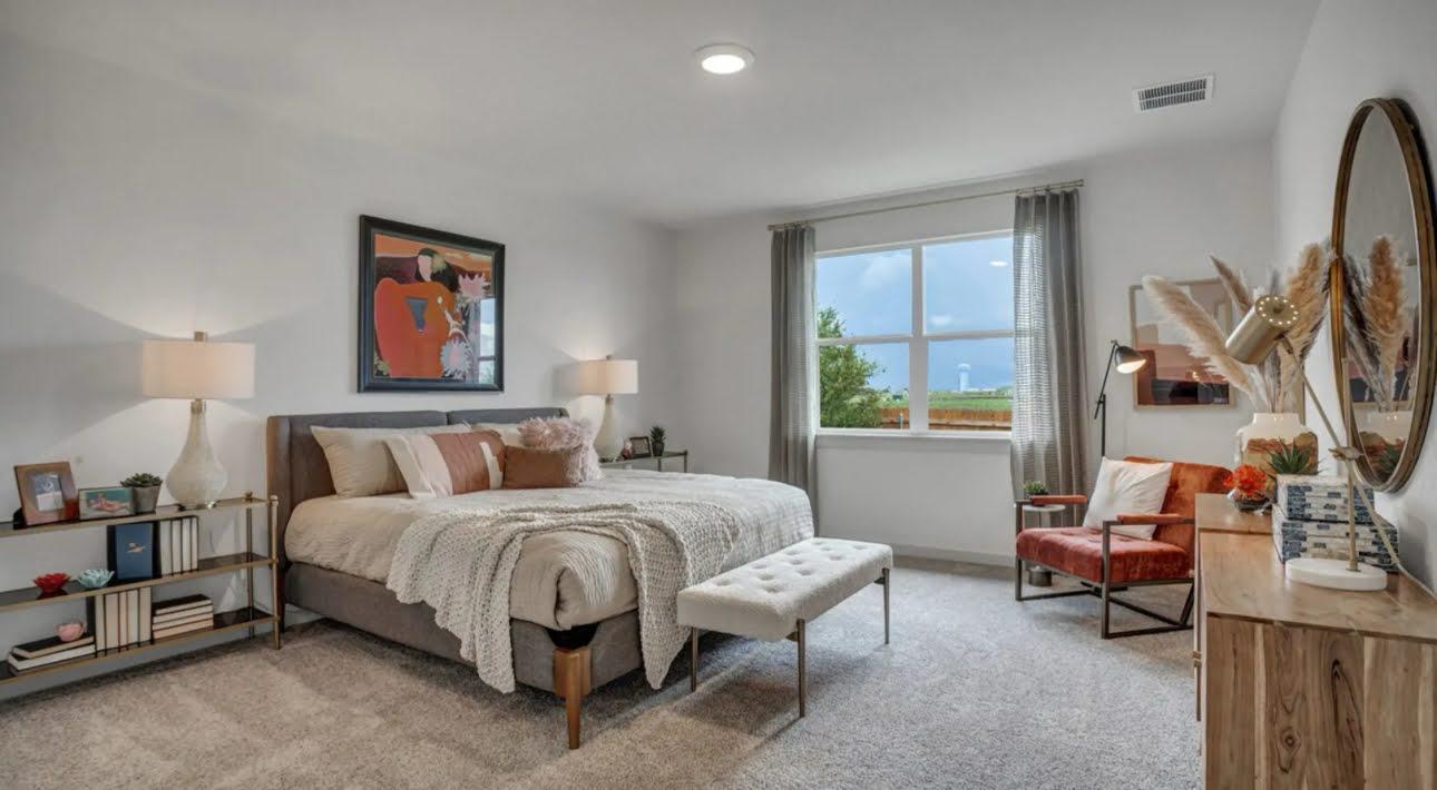 Staged bedroom with a large upholstered bed, tufted bench, and window overlooking open land.