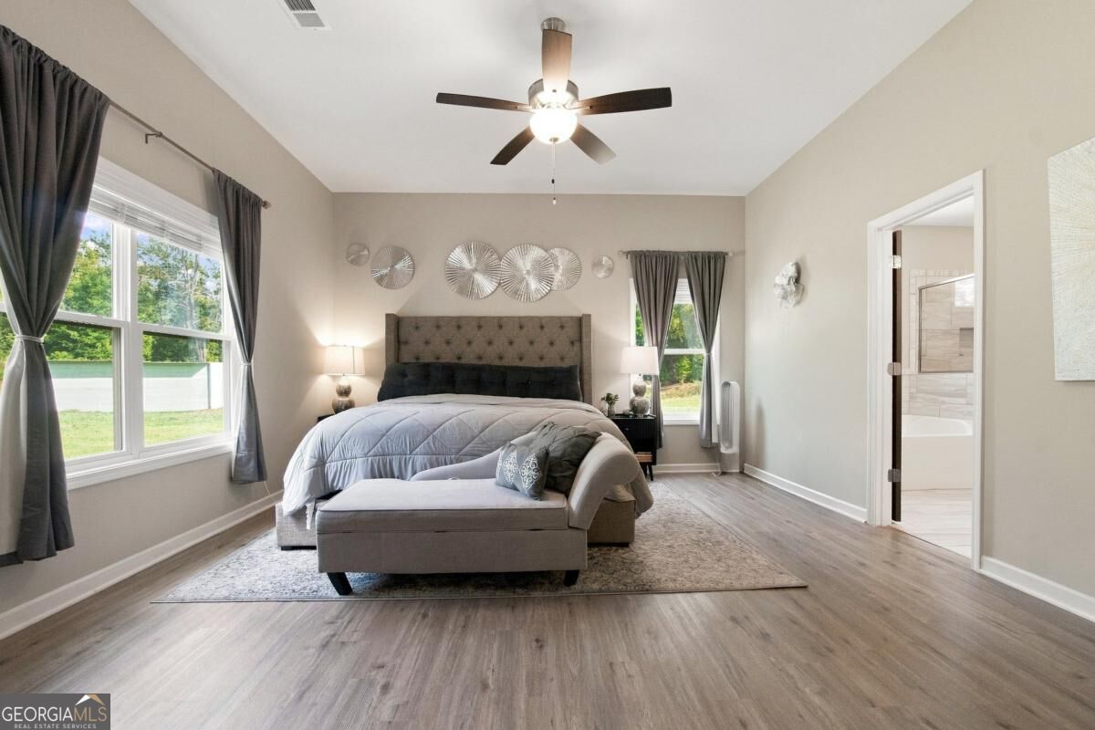 Furnished bedroom with a tufted bed, gray chaise, ceiling fan, and adjoining bathroom doorway.