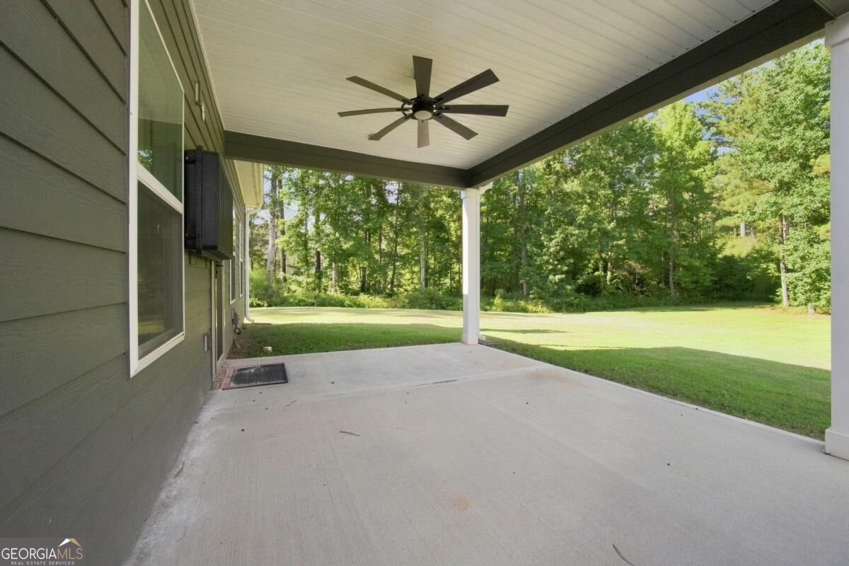 Covered concrete patio with a ceiling fan overlooking a lawn and wooded tree line.