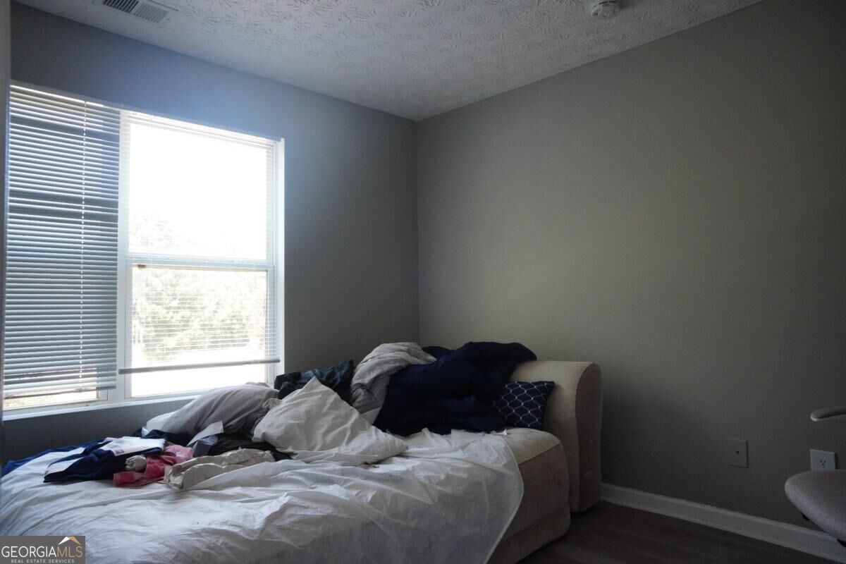Bedroom with rumpled white bedding, clothing on the bed, and a large window with blinds.