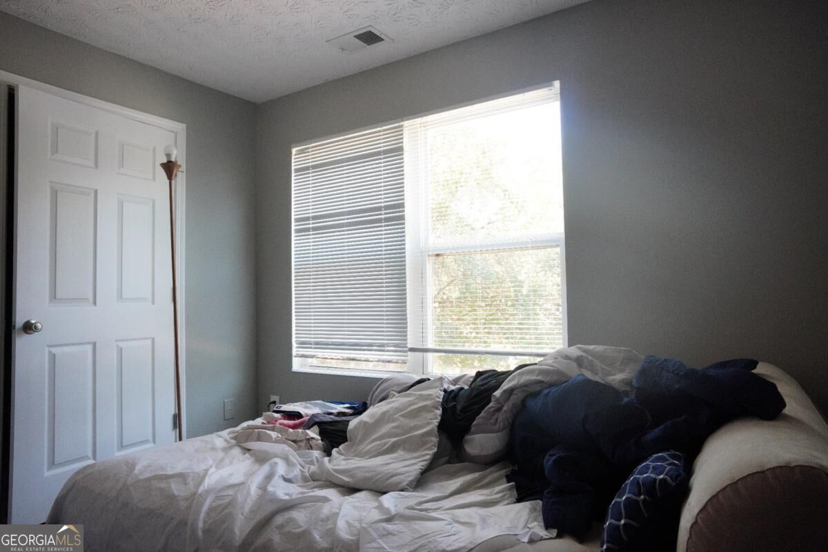 Bedroom with rumpled white bedding, dark pillows, and a large window with blinds.