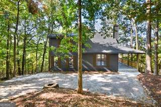Dark-sided house with a gray roof and covered porch, surrounded by trees beside a broad gravel driveway.
