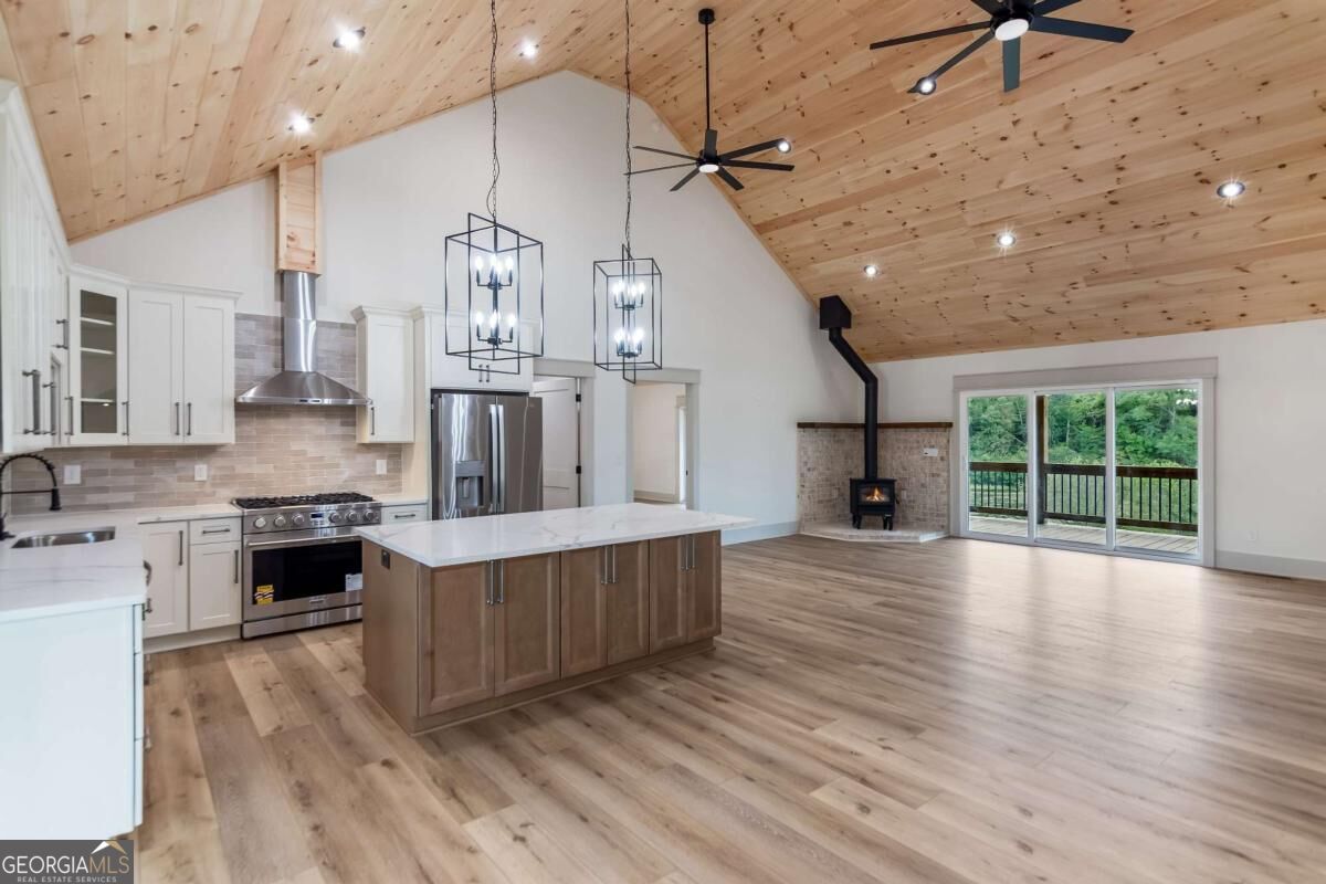 Open kitchen and living area with a vaulted pine ceiling, large island, wood stove, and wooded deck view.