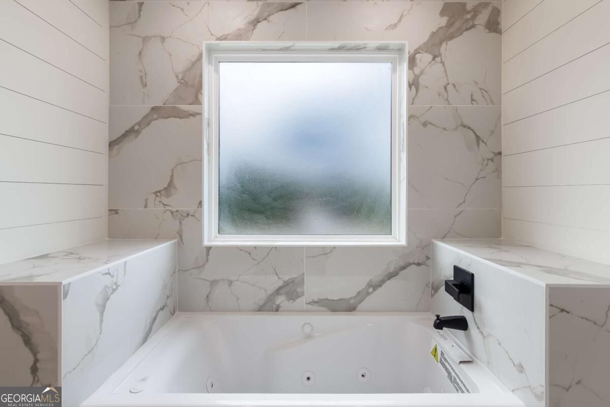 Jetted bathtub beneath a frosted window, surrounded by marble-look tile and matte black fixtures.