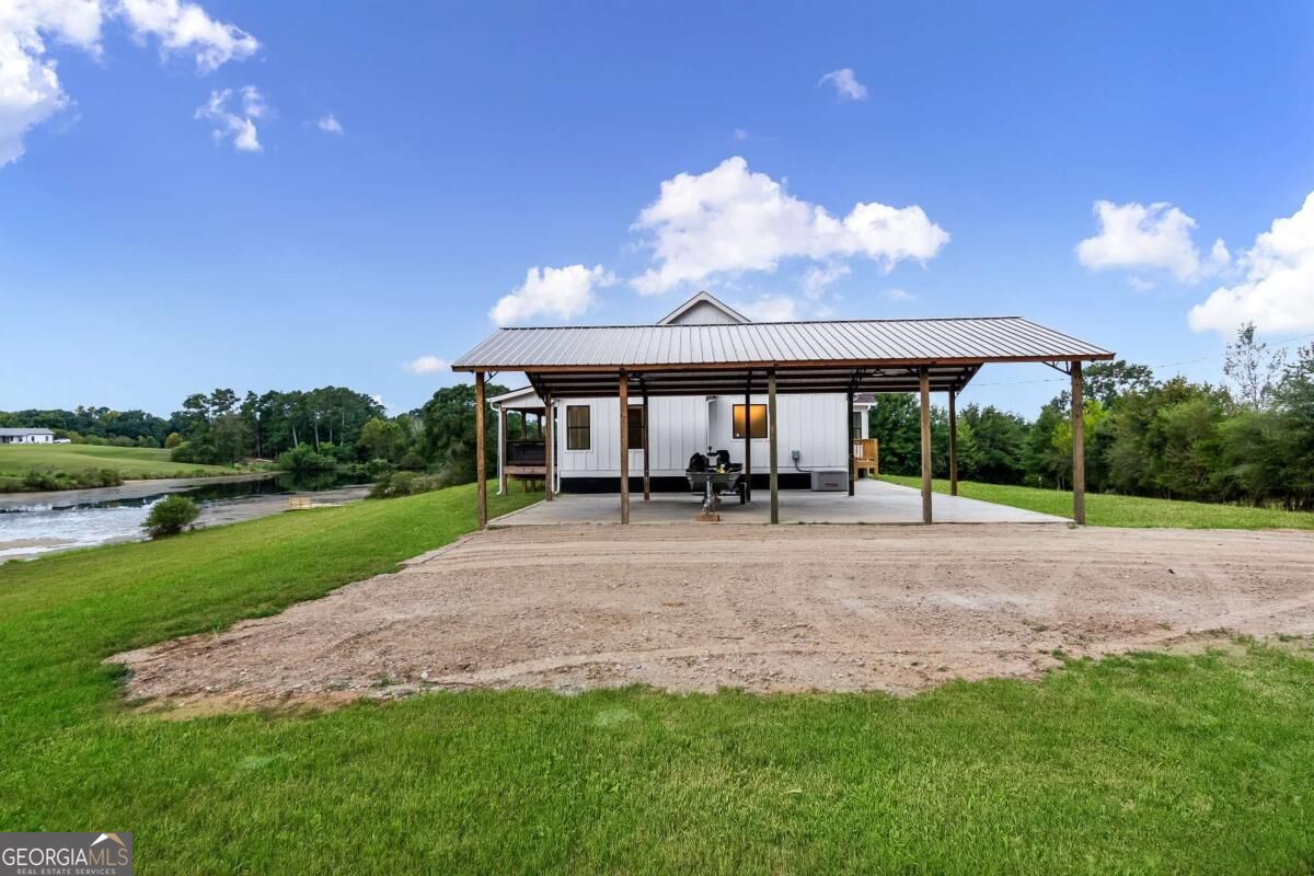 White home beneath a large covered structure beside a river and grassy yard.