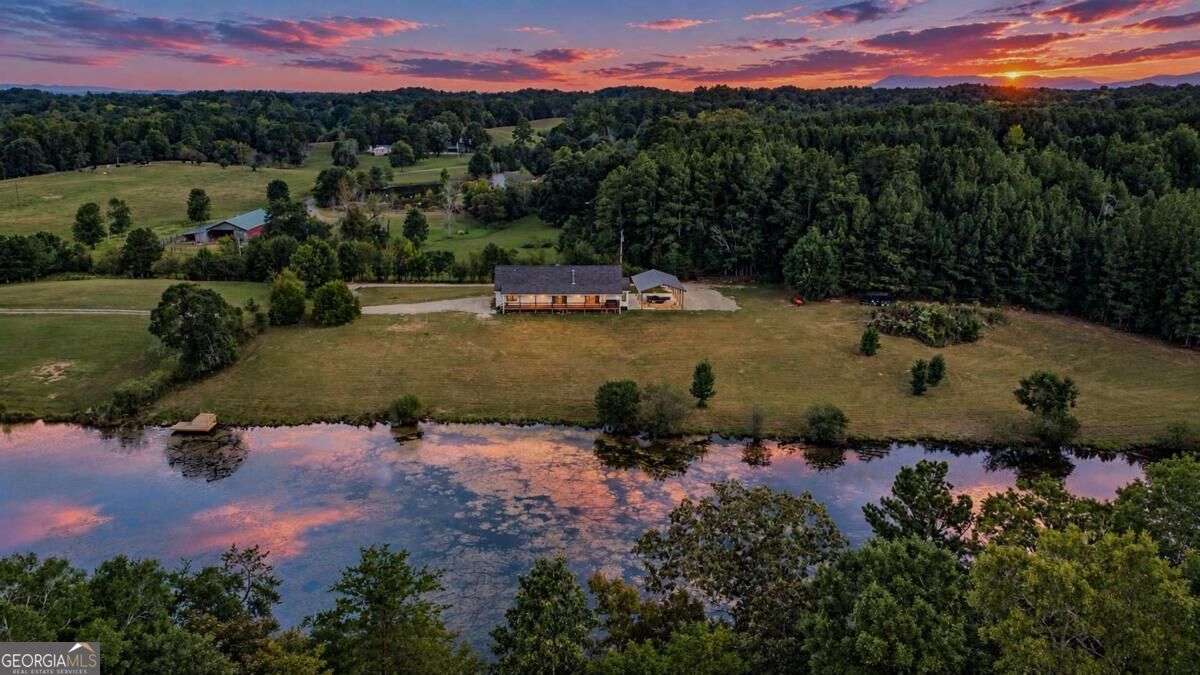 Aerial view of a home beside a pond, with wooded acreage beneath a colorful sunset.