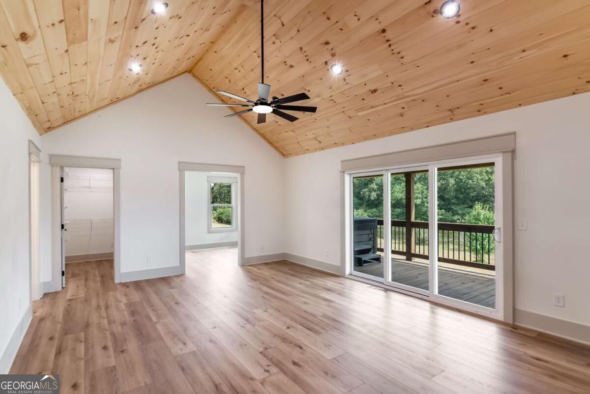 Empty living area with a vaulted wood-plank ceiling, ceiling fan, and sliding doors to a tree-lined deck.