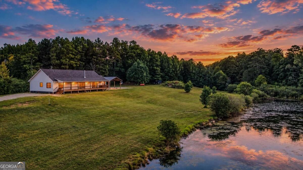 Aerial view of a home with a long covered porch beside a pond at sunset.