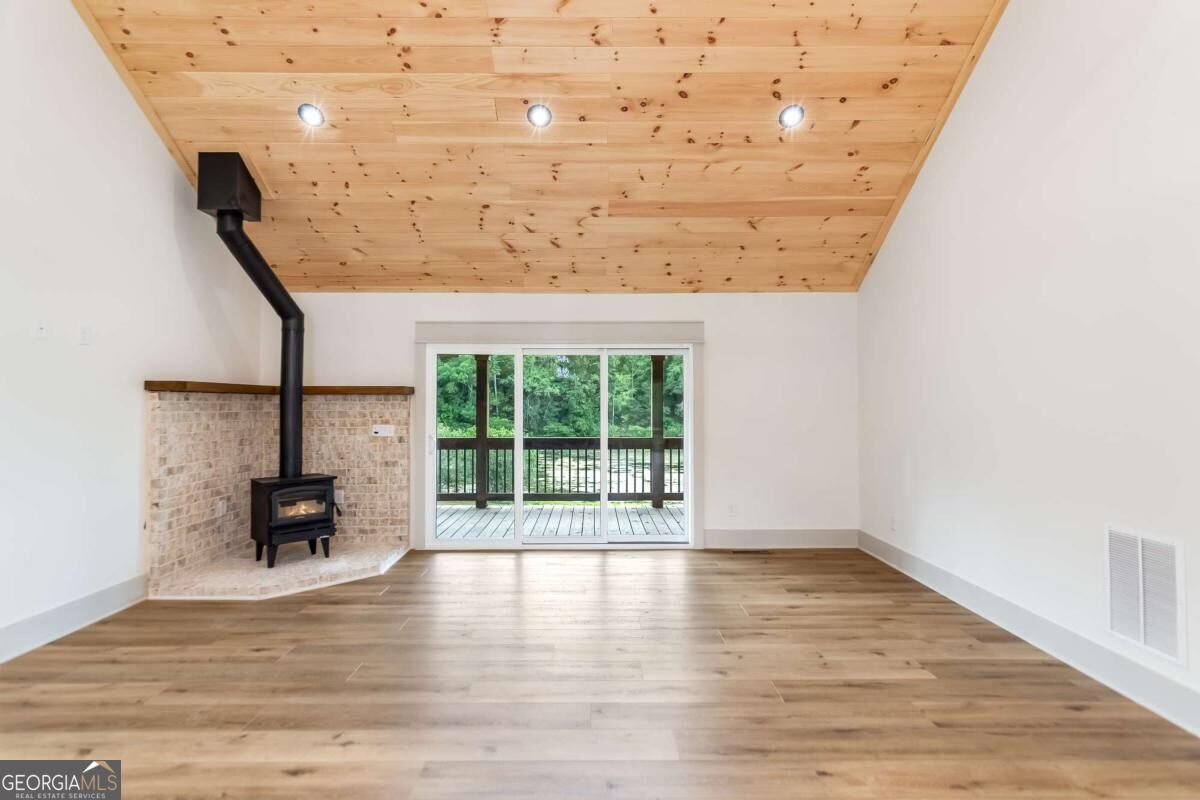 Empty living room with a vaulted pine ceiling, brick hearth, wood stove, and sliding doors to a wooded deck.