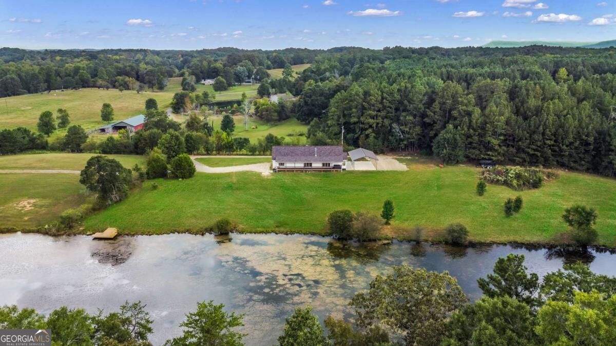 Aerial view of a rural home beside a river, with open fields and wooded hills.