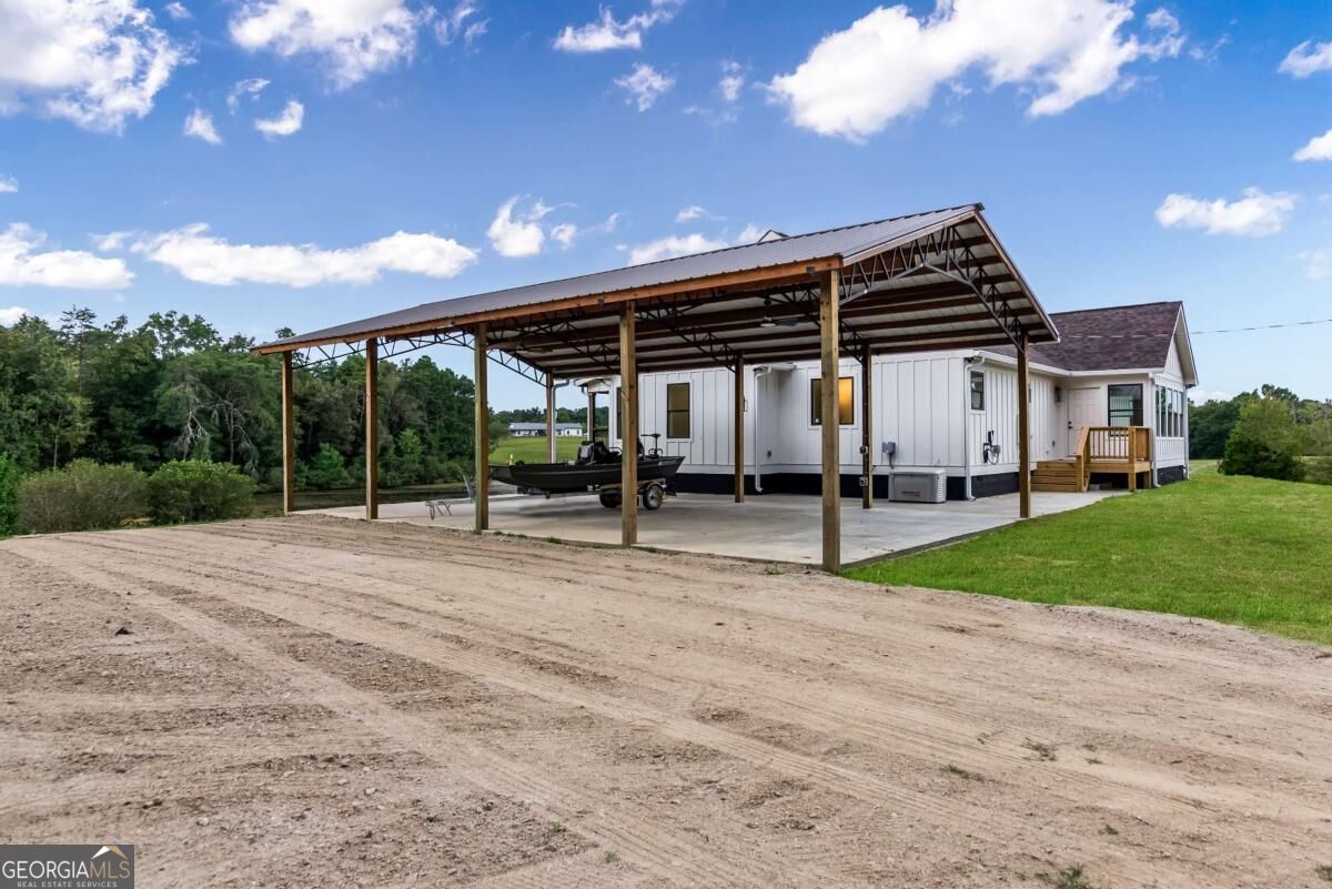 White single-story home with a large metal-roof carport, boat, and broad dirt driveway.