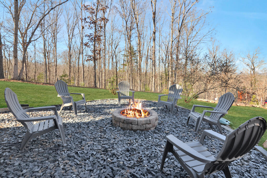 Gravel patio with a lit stone fire pit, Adirondack chairs, and leafless trees.