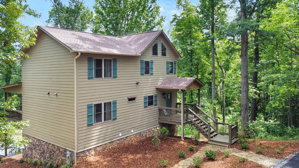 Two-story tan house with teal shutters, a covered porch, and wooden stairs among trees.