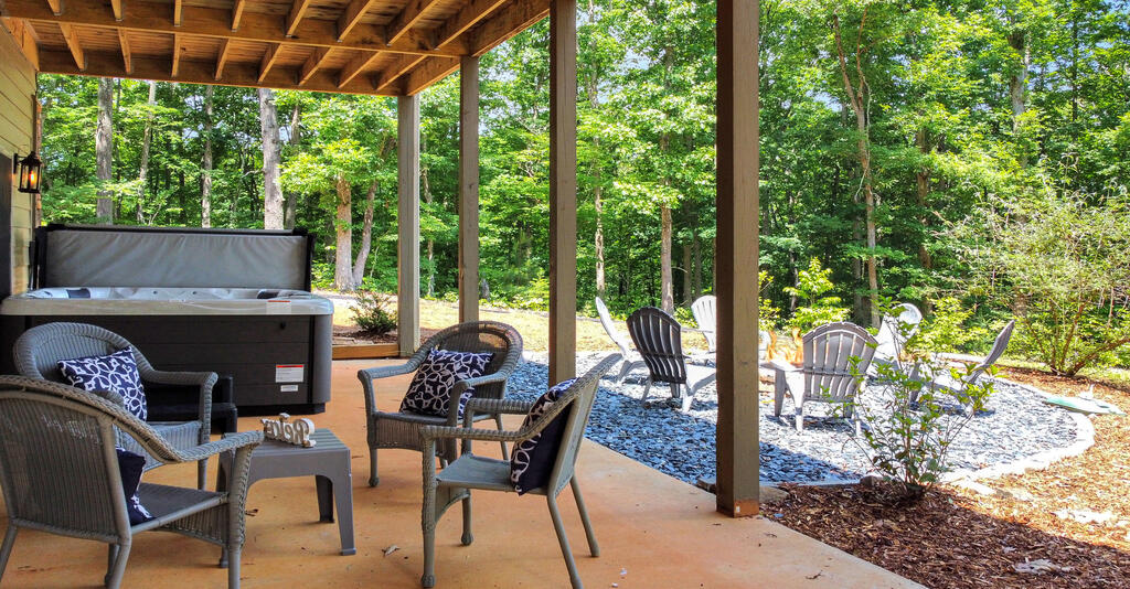 Covered patio with a hot tub and wicker seating beside a wooded stone seating area.